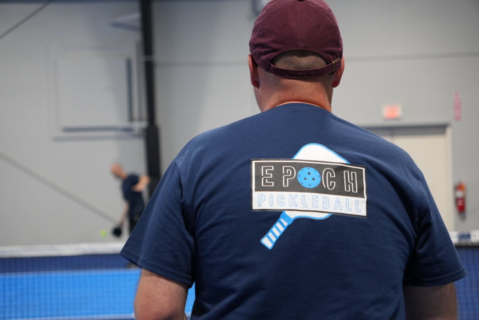 Person in maroon cap and navy shirt standing indoors at a sports court with a blue table.