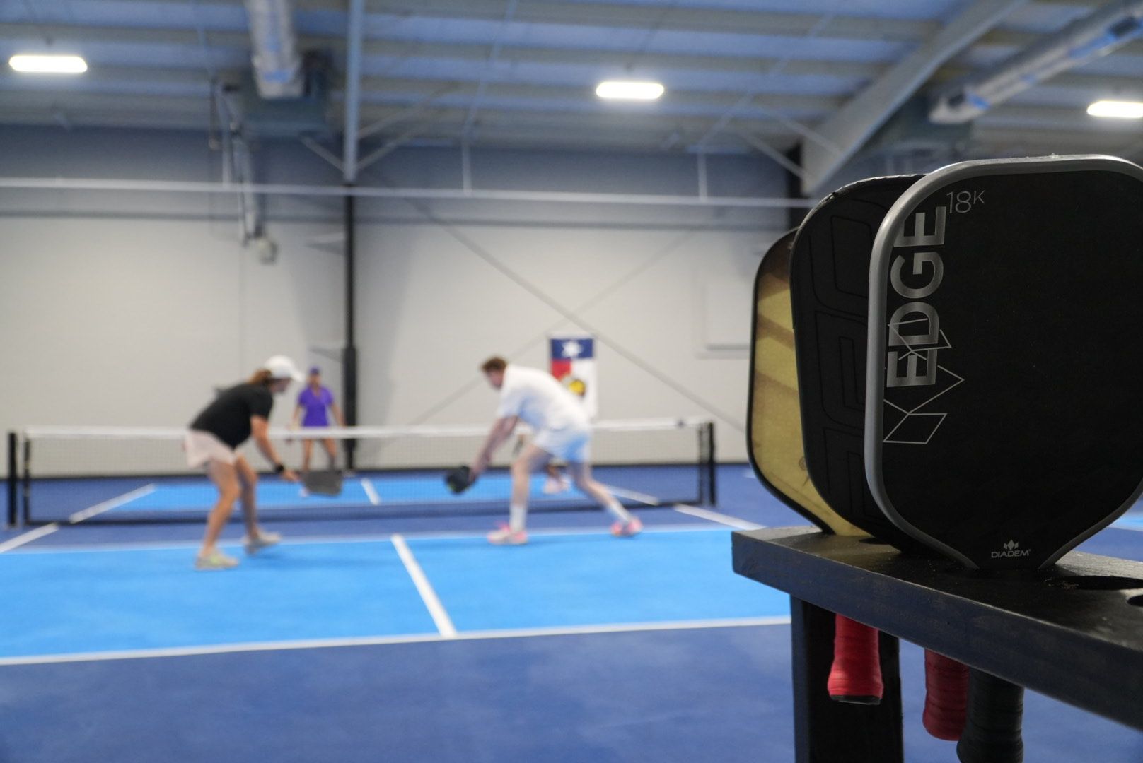 Two pickleball players in action on a blue indoor court, with a black paddle in the foreground.