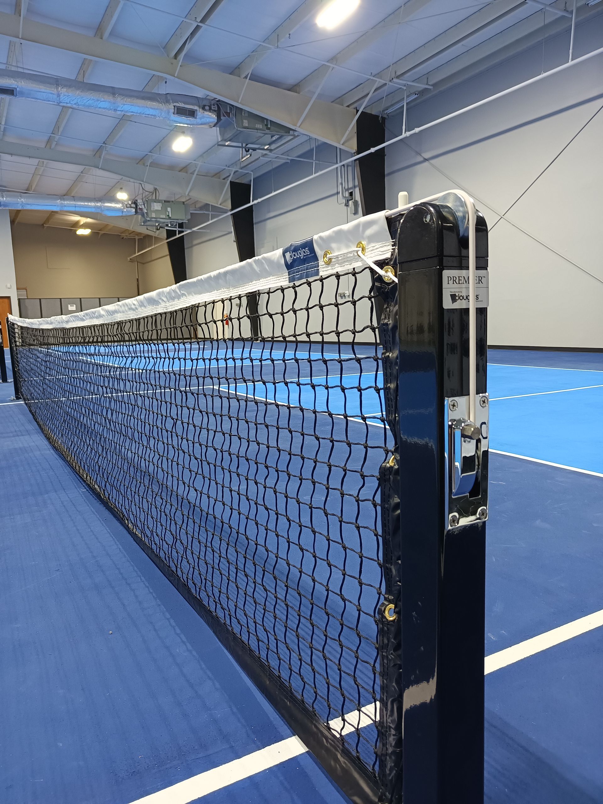 Close-up of a black tennis net on an indoor blue court with white lines and ceiling lights