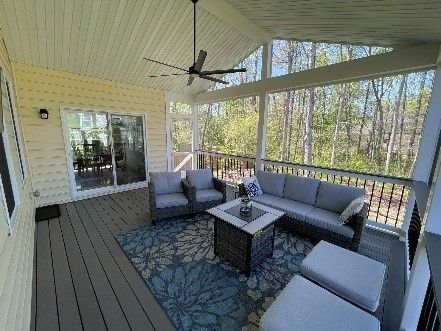 Covered porch with gray sectional seating, blue rug, ceiling fan, and wooded view beyond railing