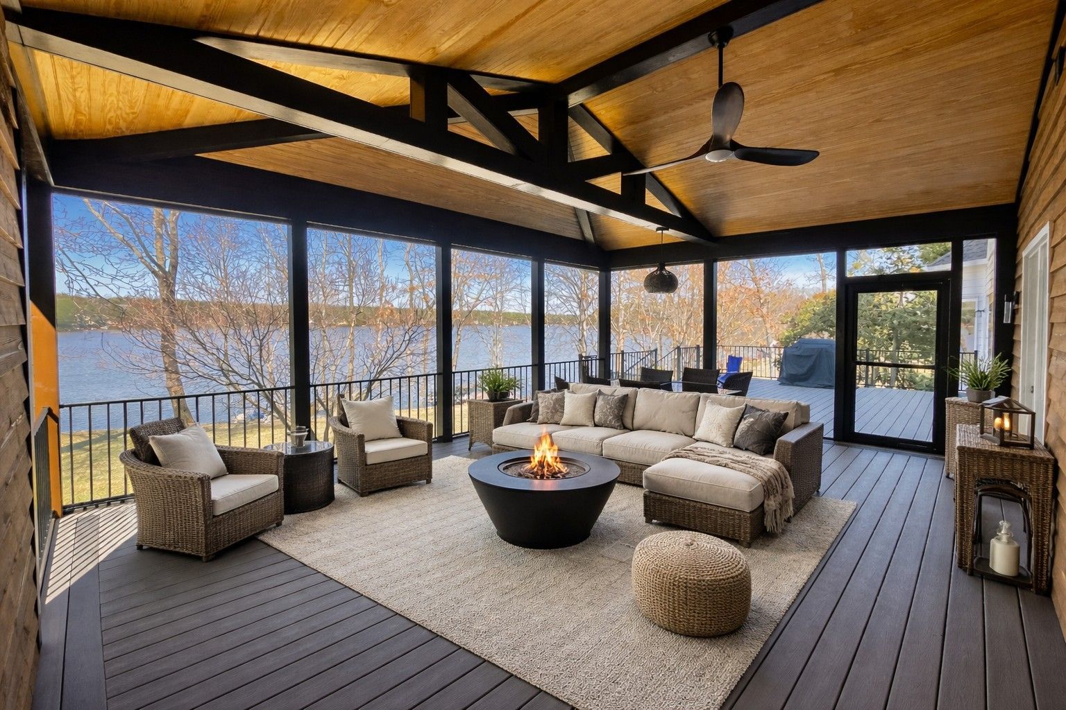 Covered outdoor patio with wicker seating, fire pit, and dark wood ceiling overlooking a lake