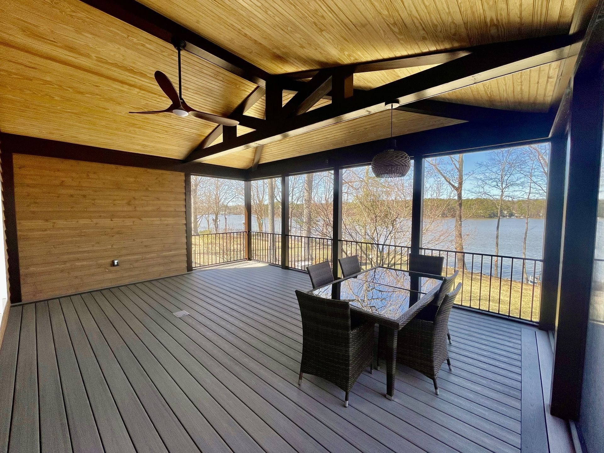 Covered wooden deck with dining table and chairs, overlooking a bright outdoor view through glass railings.