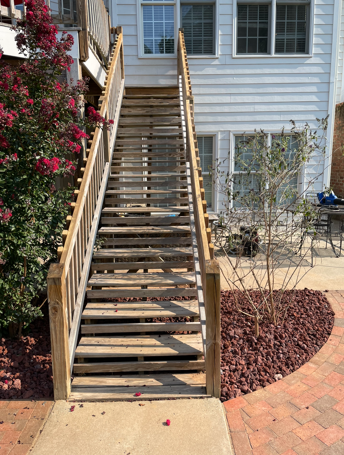 Wooden outdoor staircase leading to a white house, with a flower bed and brick patio below