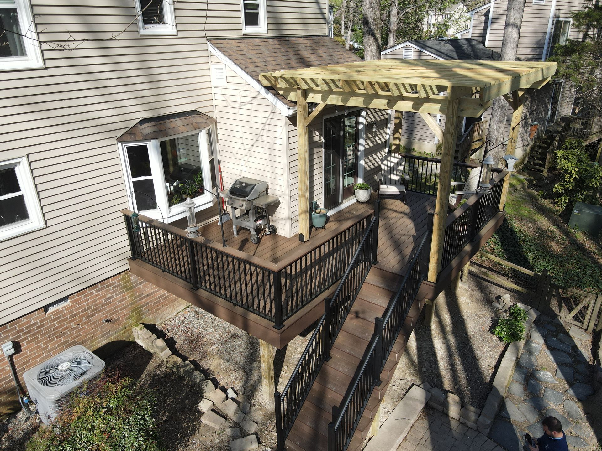 Back deck with black railing, pergola, patio chairs, and grill beside a beige house