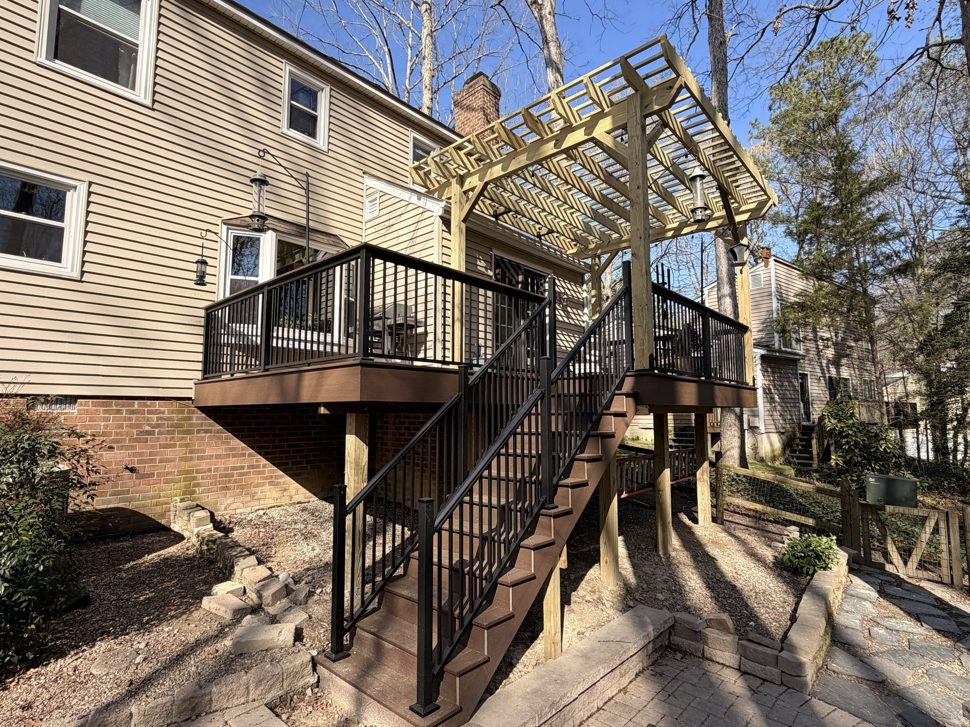 Back deck with black railings and stairs, supported by posts, beside a beige house and gravel yard.