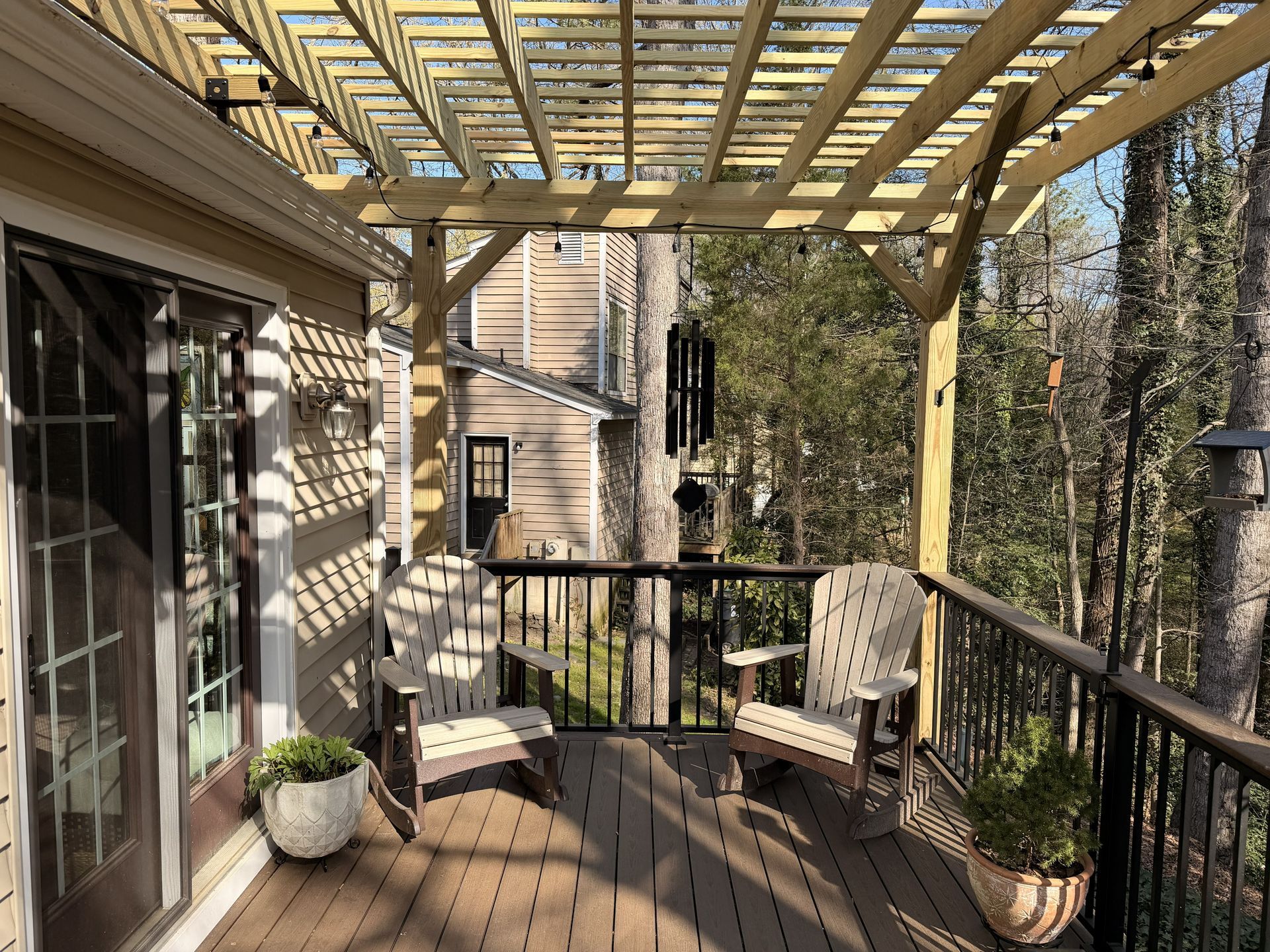 Wooden deck with pergola, sliding glass door, railing, potted plants, and two chairs overlooking trees