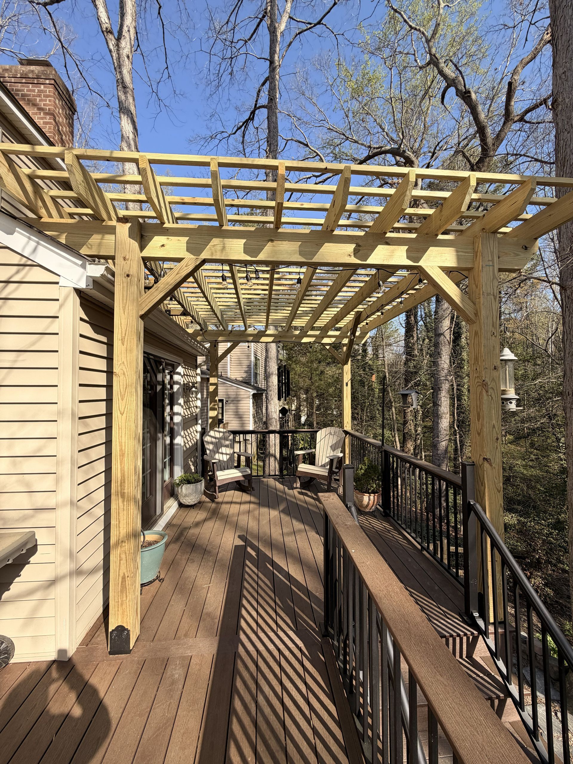Wooden deck with pergola framing a narrow walkway beside a tan house, surrounded by trees.