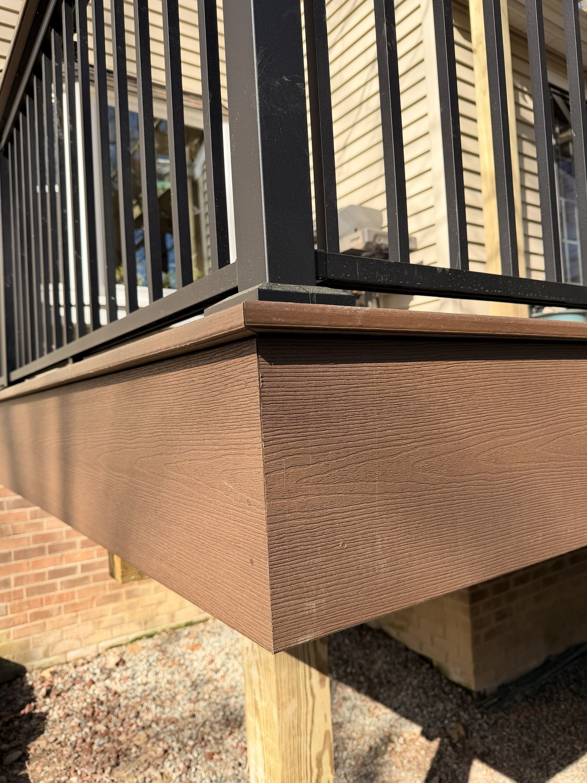 Brown deck corner with black metal railing and textured skirting above a brick wall and concrete support post