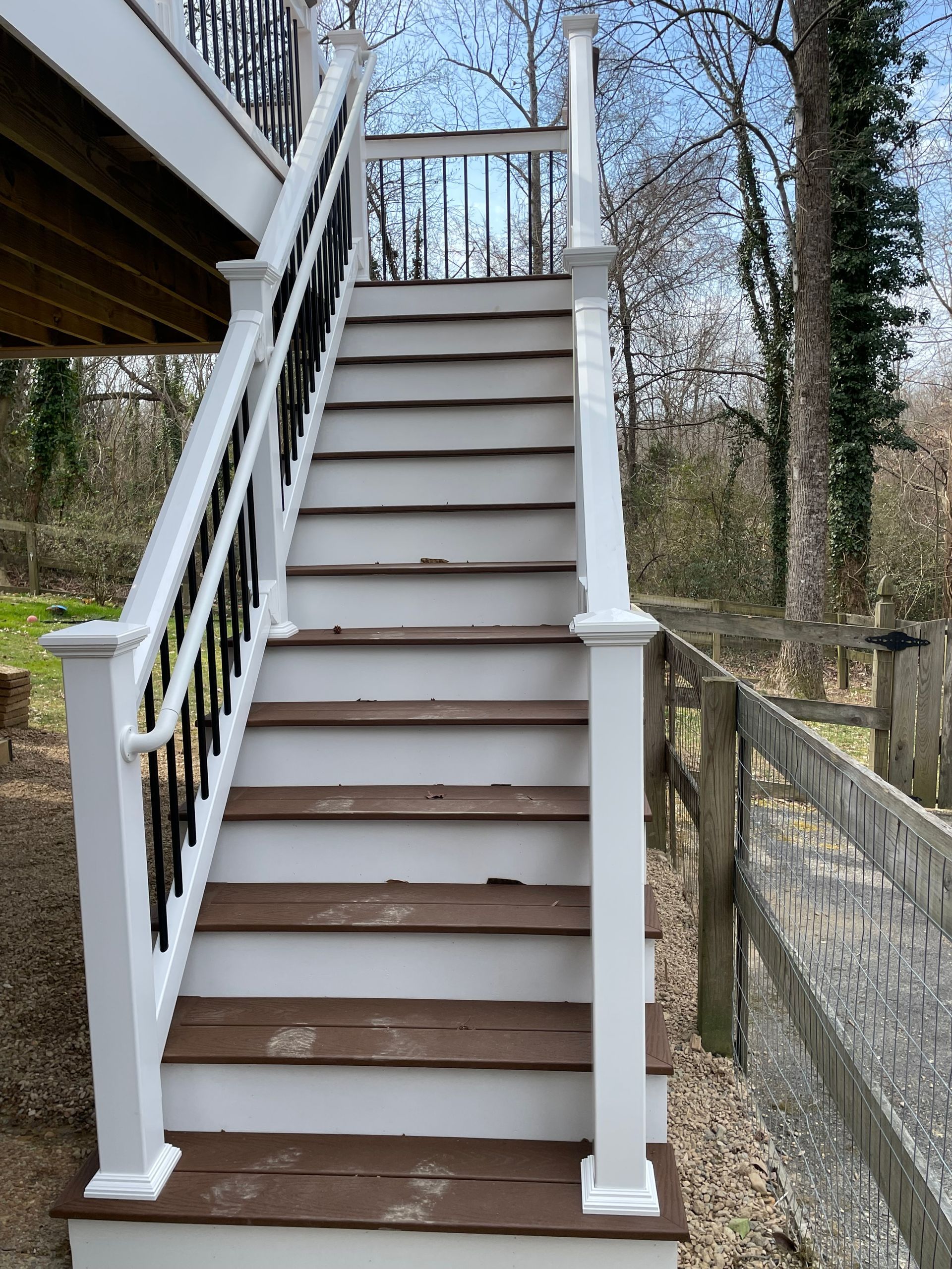 Outdoor wooden staircase with white railings leading up to a deck, beside a stone wall and trees