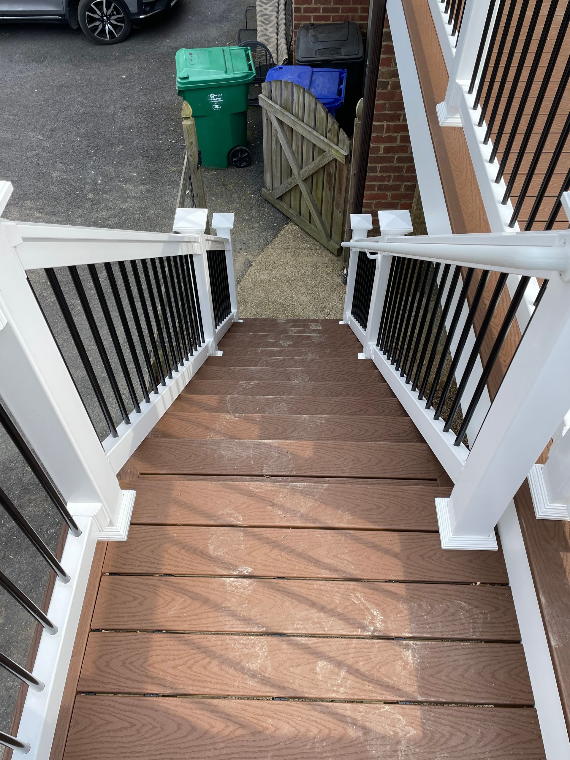 Front steps with brown treads, white railings, and a small landing leading to a narrow path by bins and a fence.