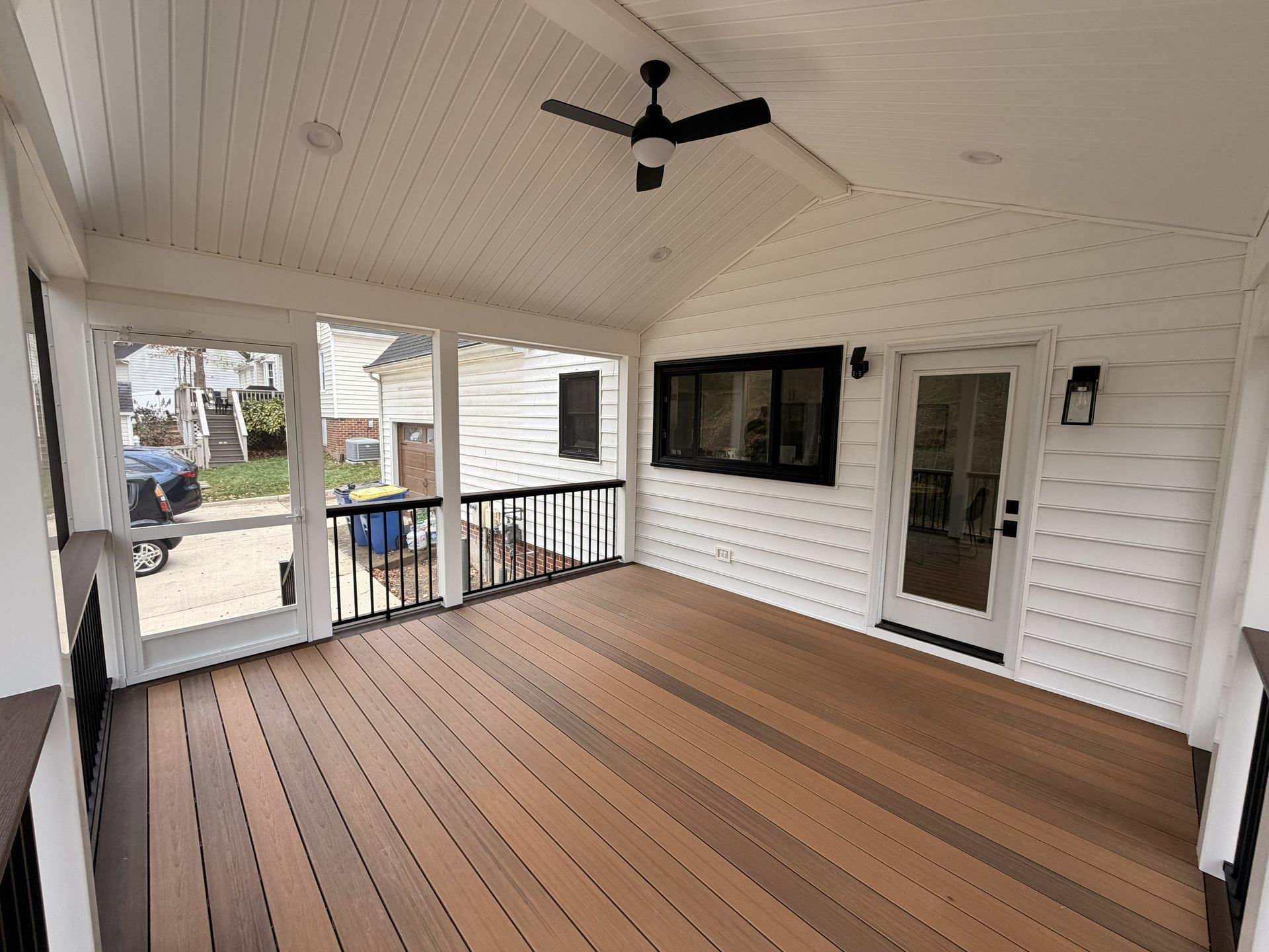 Covered front porch with wood-look flooring, white siding, black ceiling fan, railing, and glass front door.