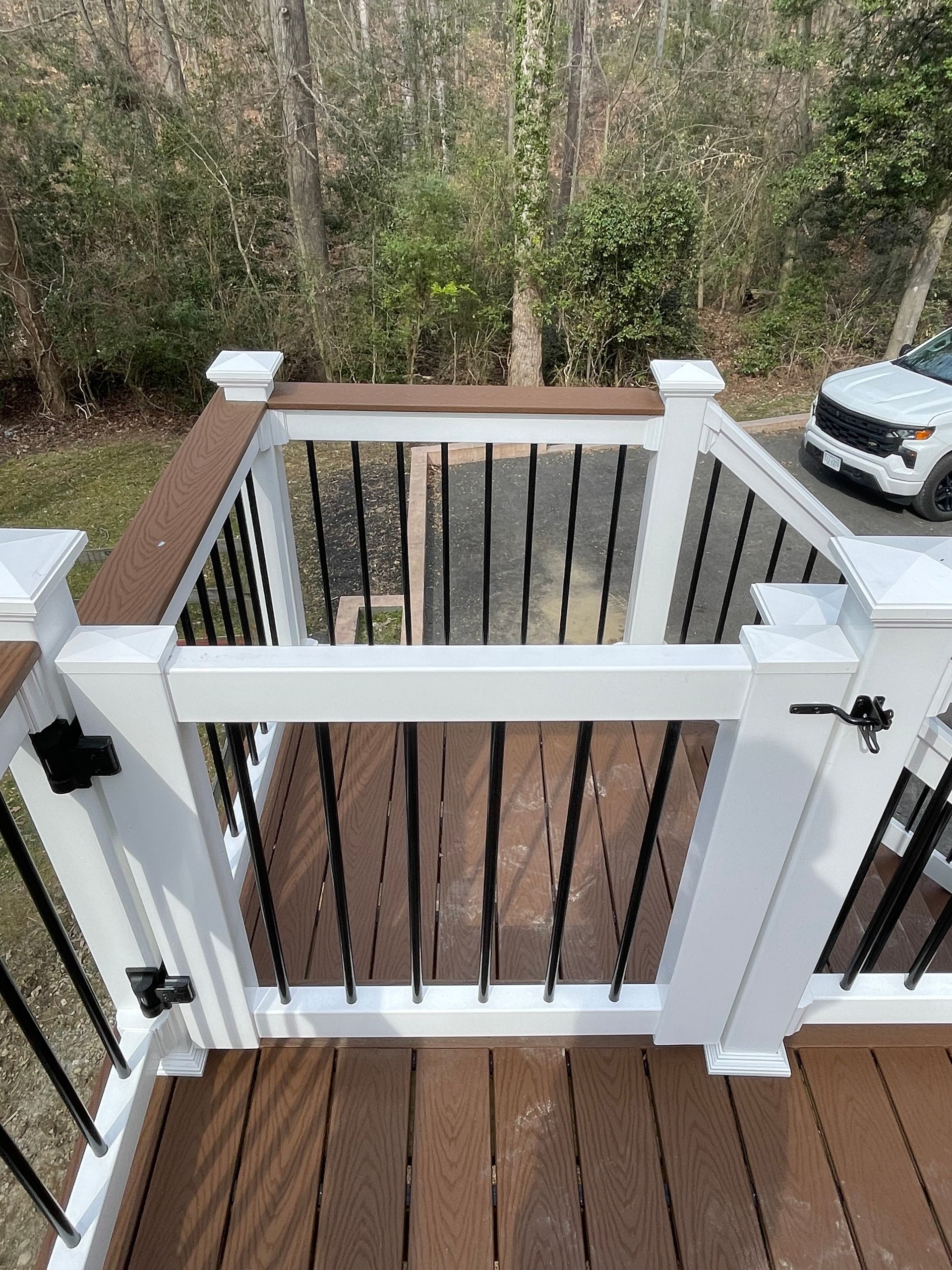 White deck gate with black railing overlooking a wooded yard and a parked car below.