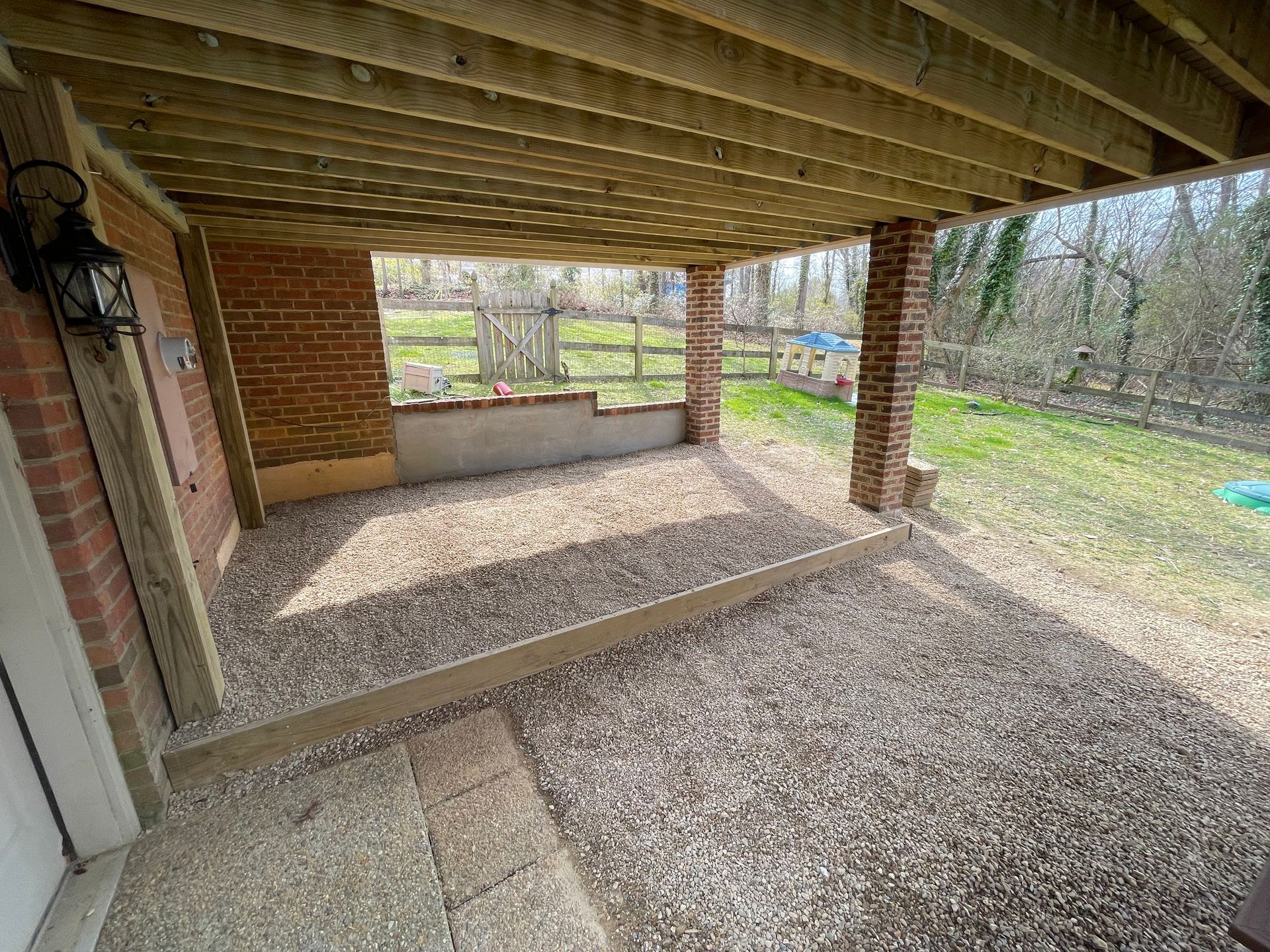 Covered brick patio with wooden beams, gravel floor, and a view of a yard and trees beyond