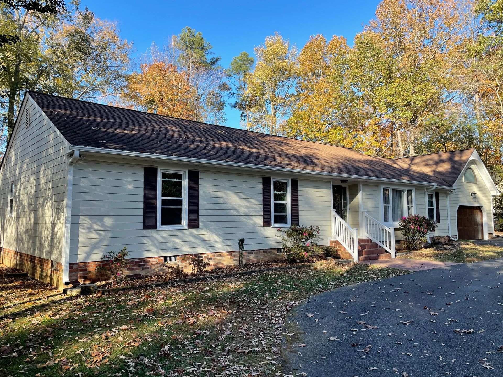 White house with dark roof and white shutters beside a driveway, surrounded by autumn trees.