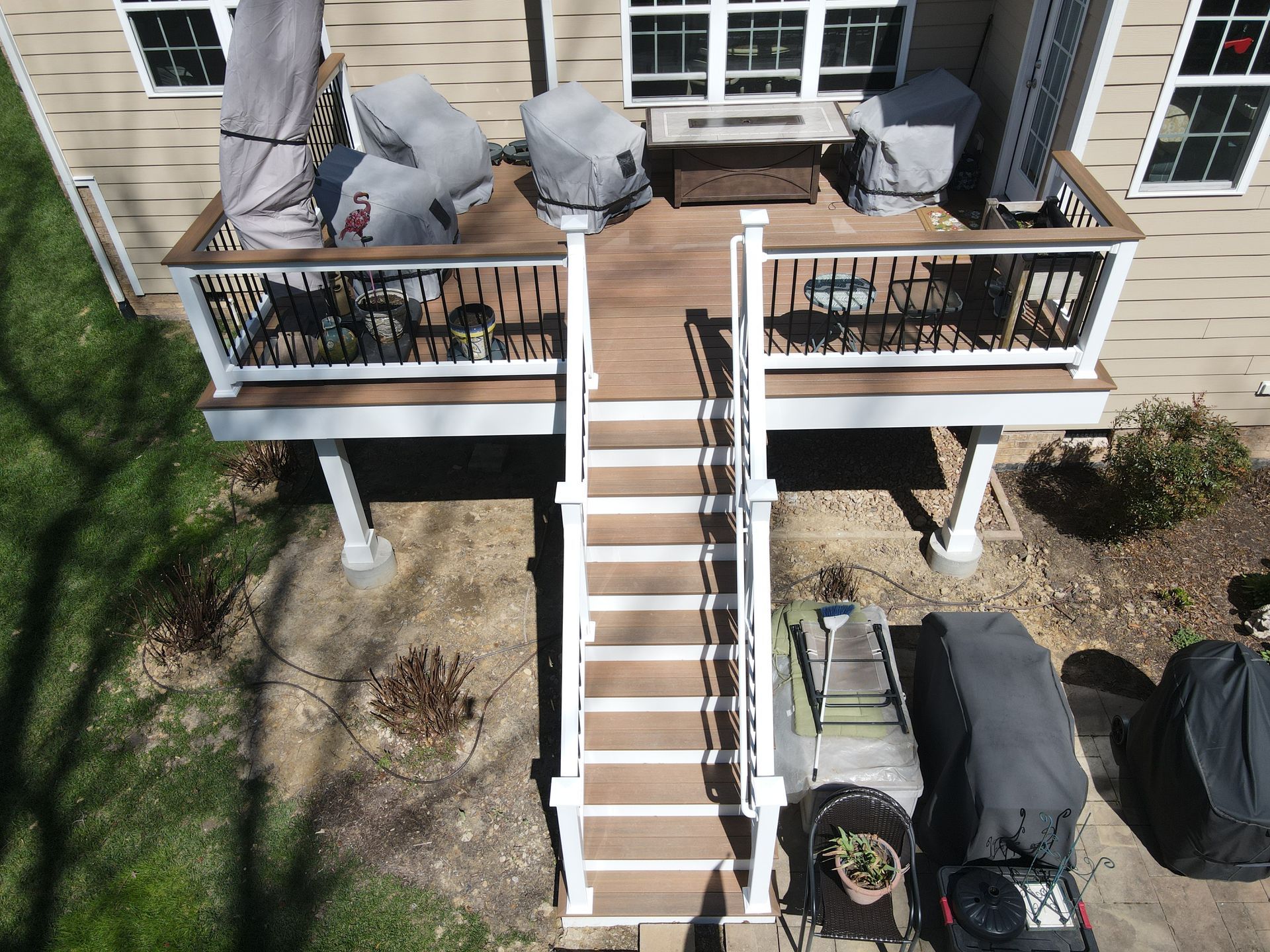 Aerial view of a house deck with a central staircase, patio furniture, and covered grills in the yard.