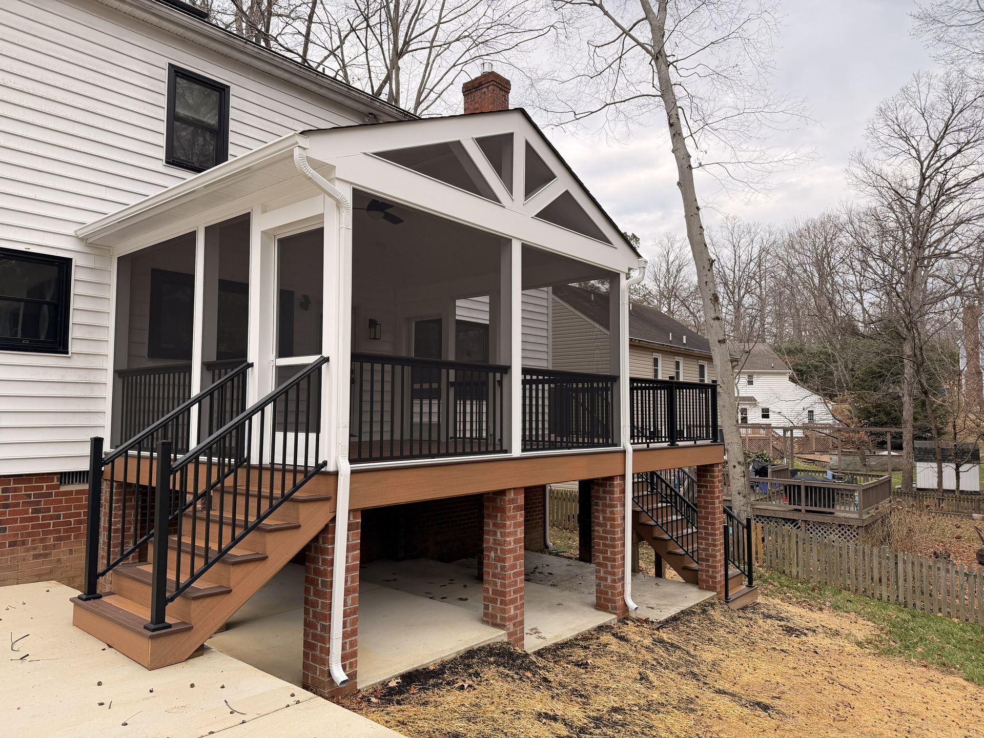 Covered wooden porch with black railings and stairs on a white house with brick foundation.