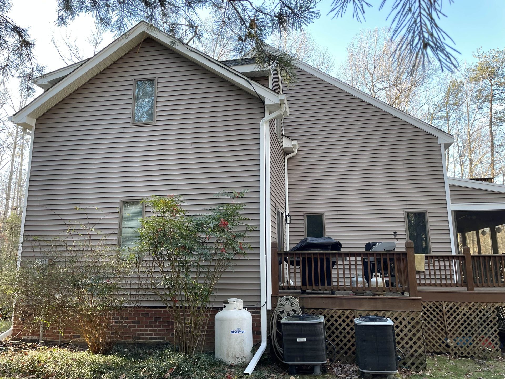 Side view of a beige house with a small back deck, grill, and trash bins in a wooded yard.