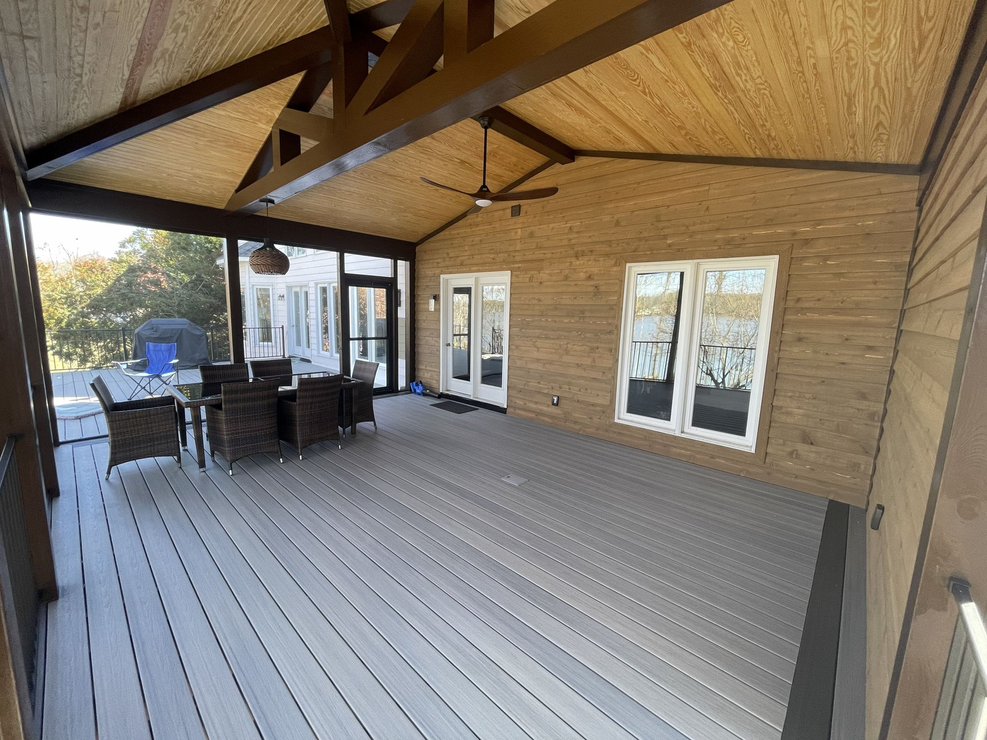 Covered wooden patio with dining table, chairs, and sliding glass doors beside a house.