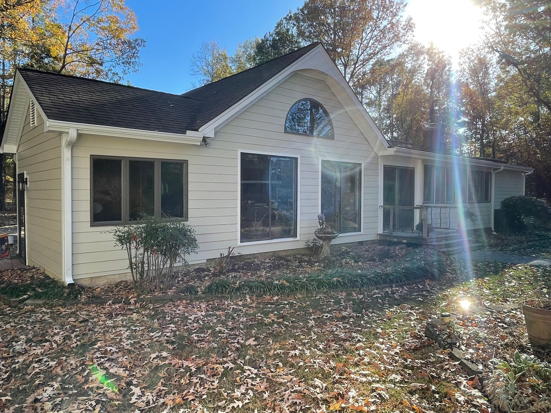 Single-story cream house with dark roof and autumn leaves in the yard, sun shining through trees