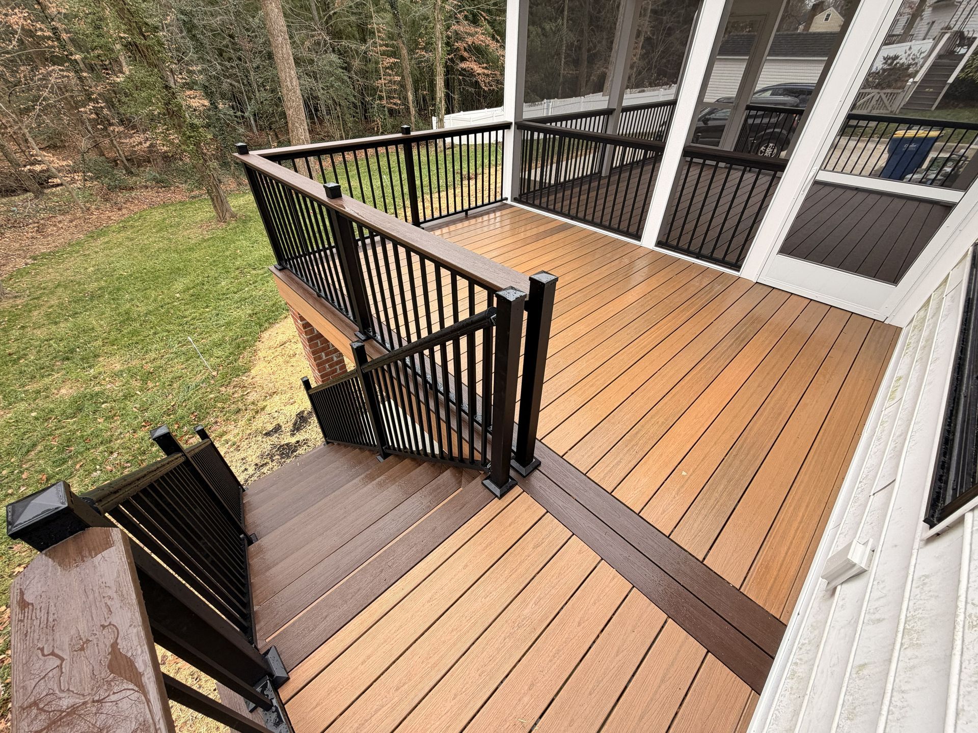 Wooden deck with black railings and stairs beside a house, overlooking a grassy yard and trees