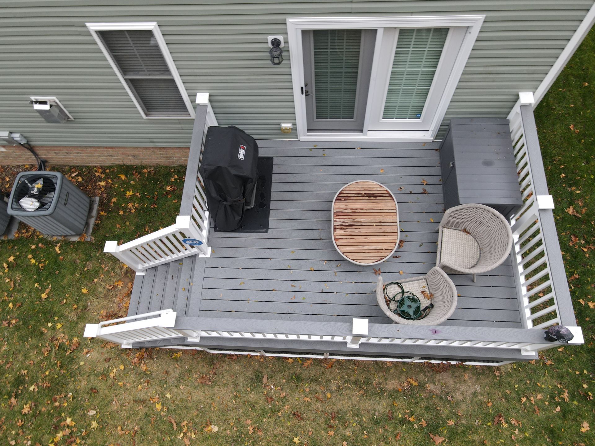 Overhead view of a small gray deck with patio chairs, table, grill, and sliding door beside a yard