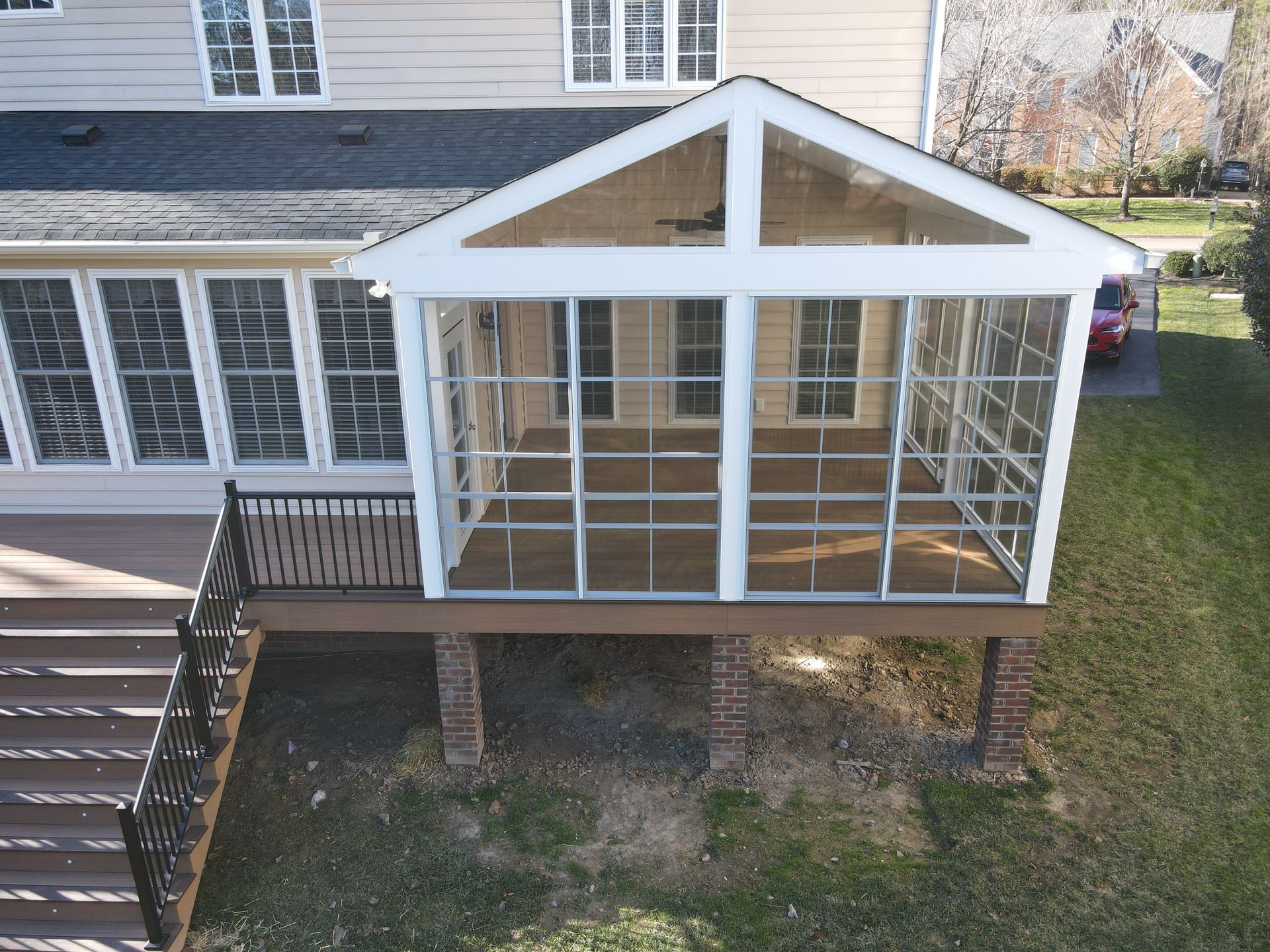 White screened porch with a gabled roof and black railing attached to a house, viewed from the backyard