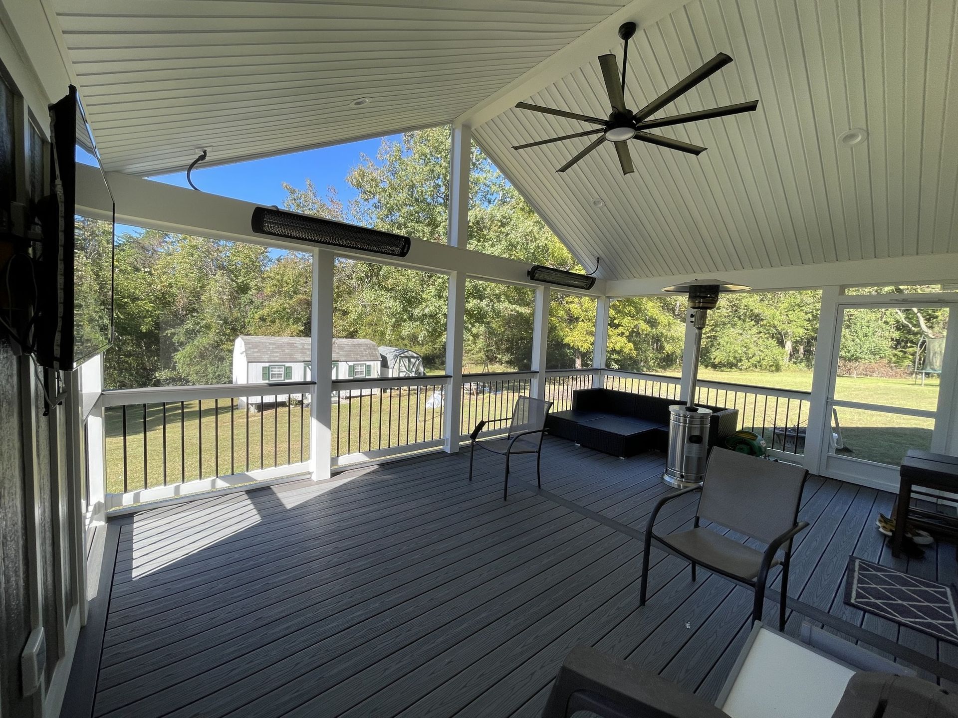 Covered porch with gray decking, black ceiling fan, railing, and chairs overlooking a green yard.