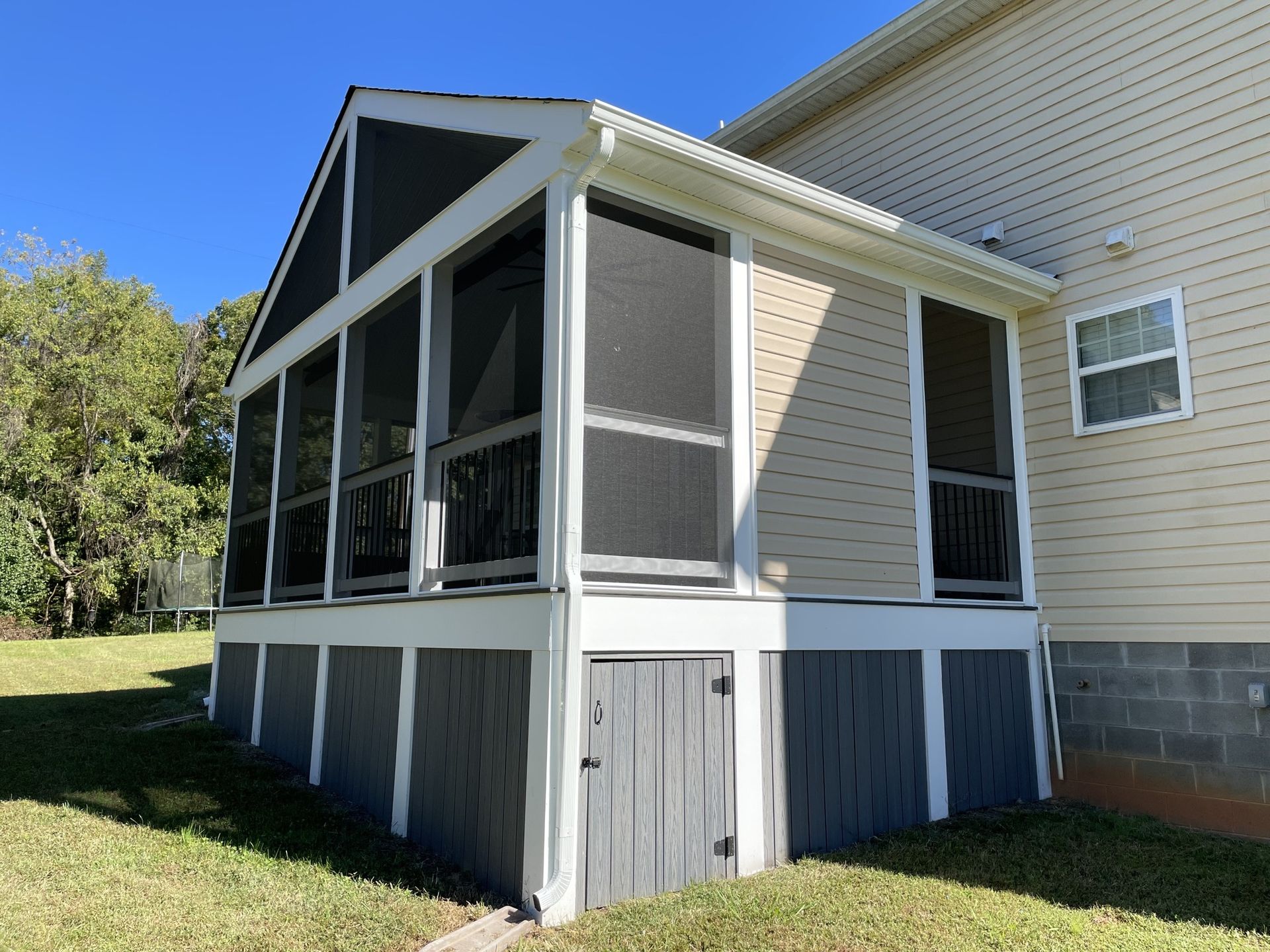 Screened porch attached to a beige house with white trim and blue-gray skirting, beside a grassy yard.