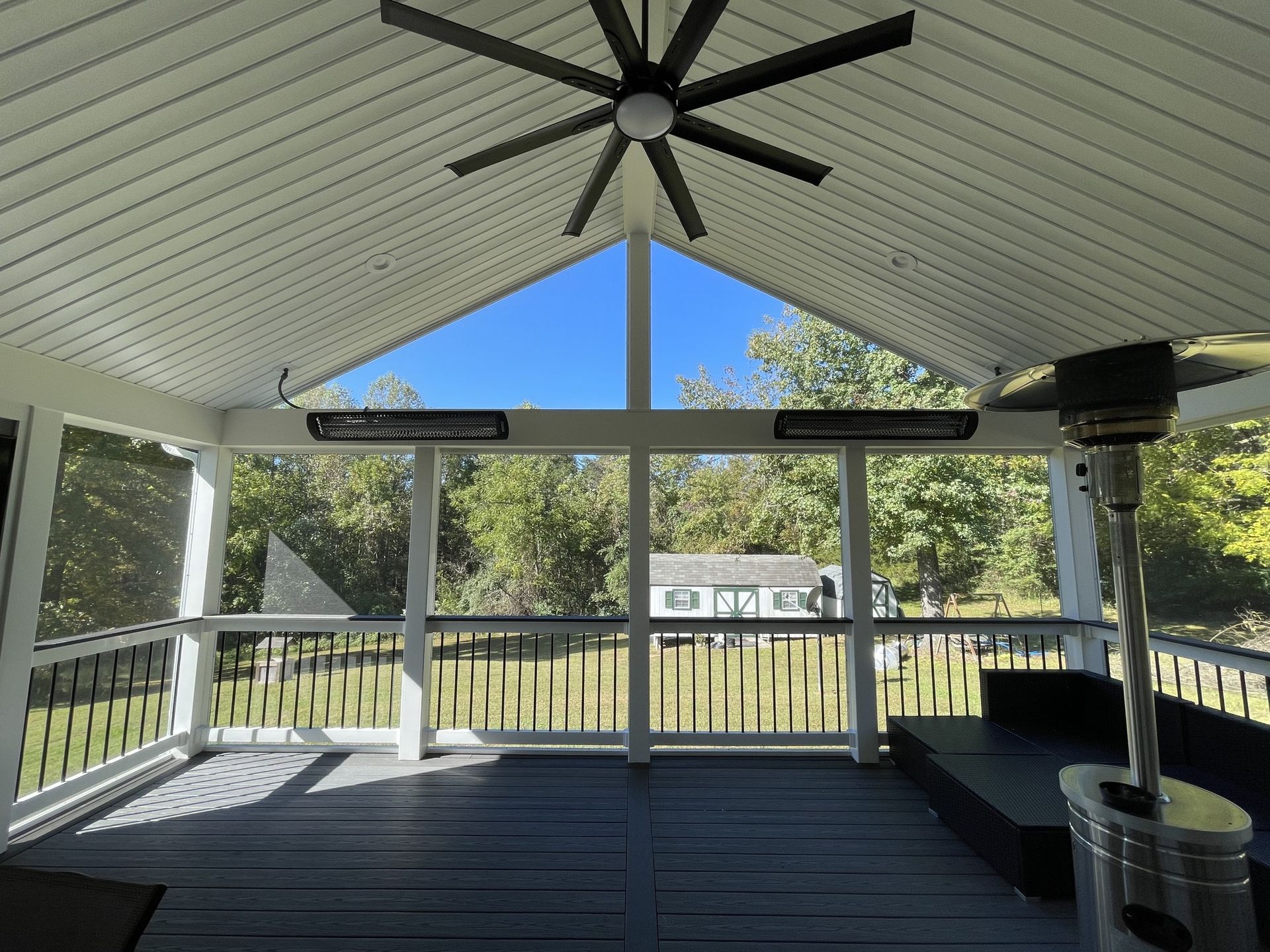 Covered porch with white railing, dark floor, ceiling fan, and trees visible through large windows.