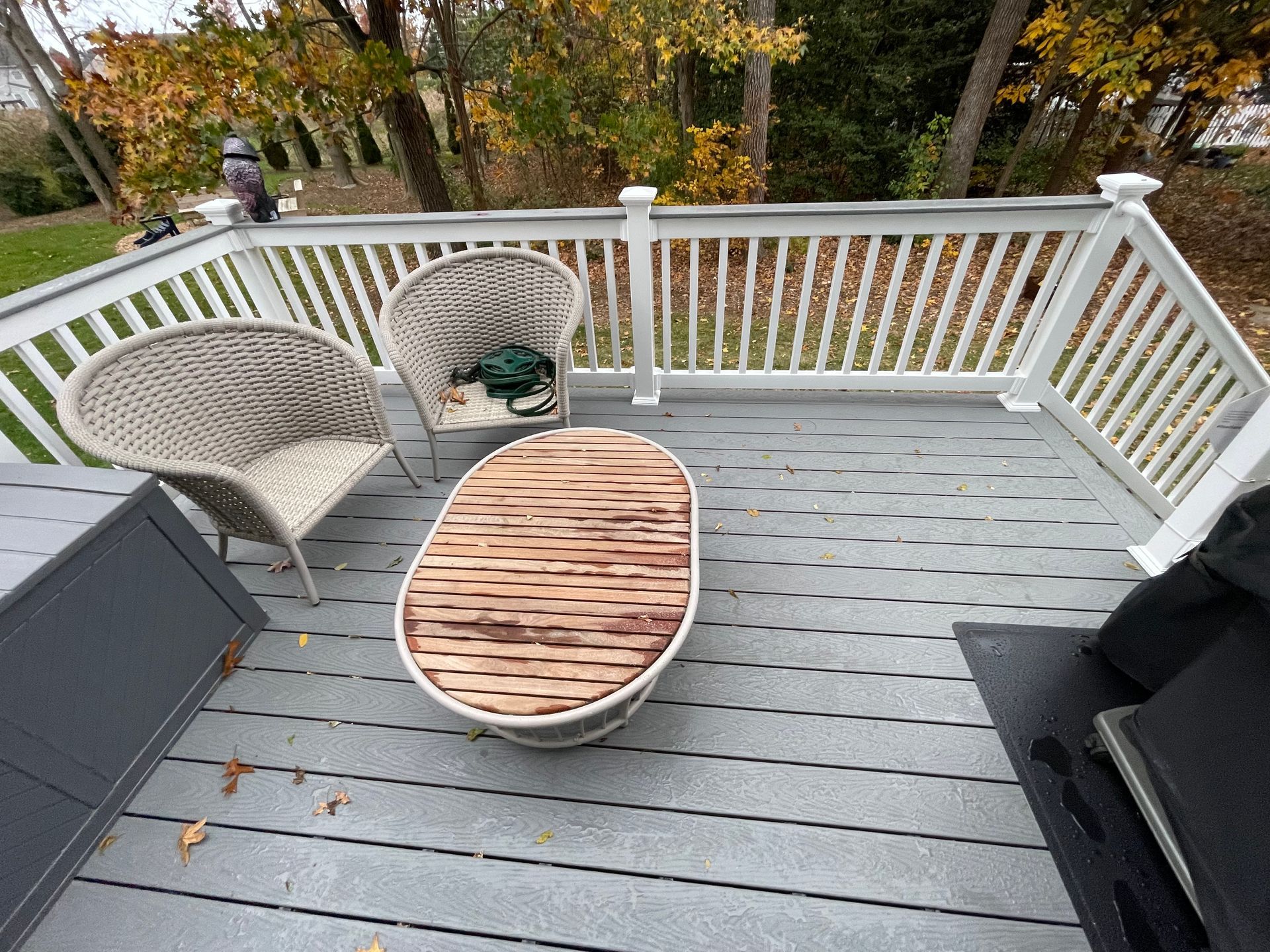 Deck with wicker chairs, oval wooden table, and railing overlooking an autumn yard.