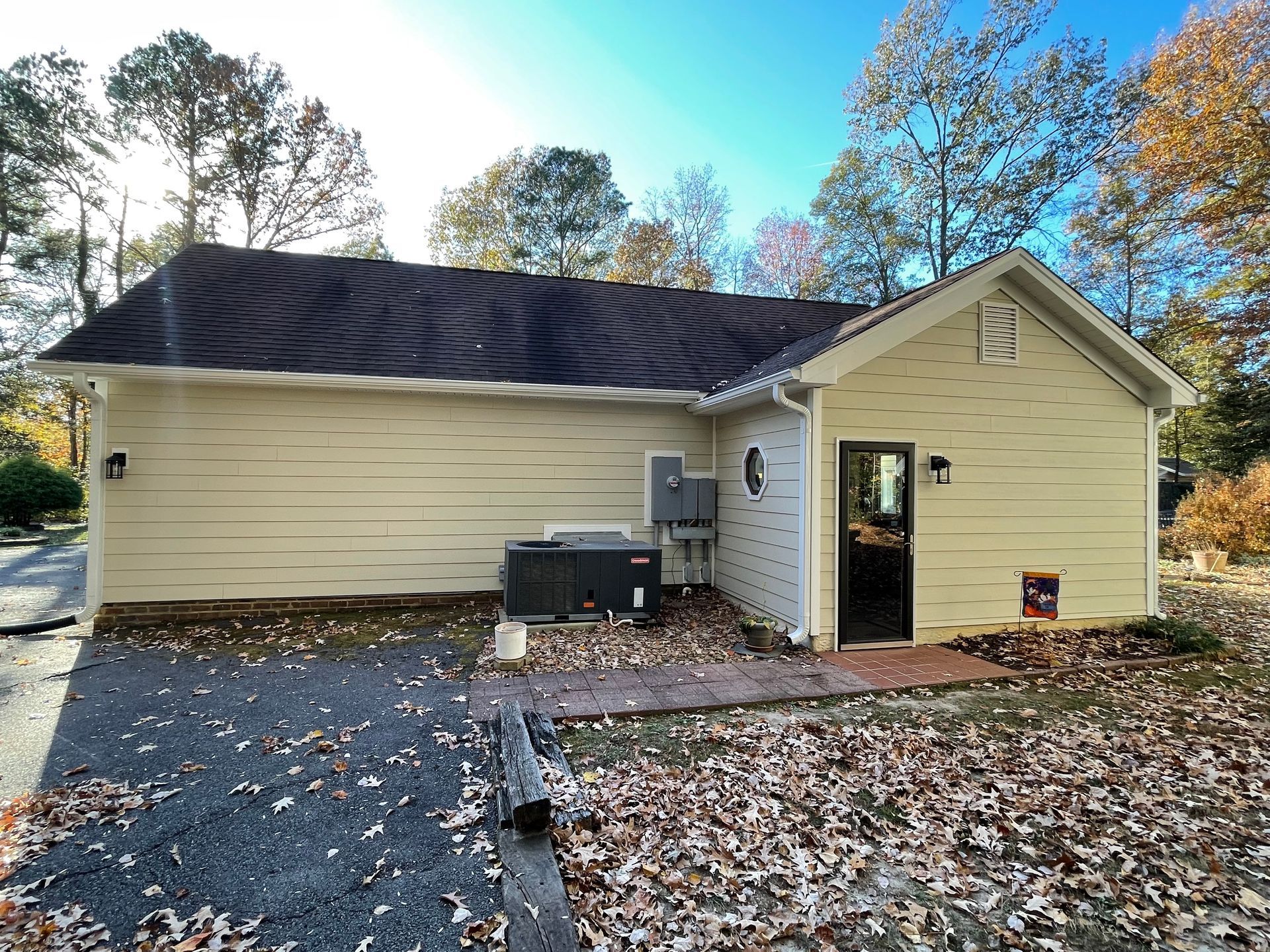 Small beige house with dark roof, open side door, outdoor AC unit, and fallen leaves in the yard
