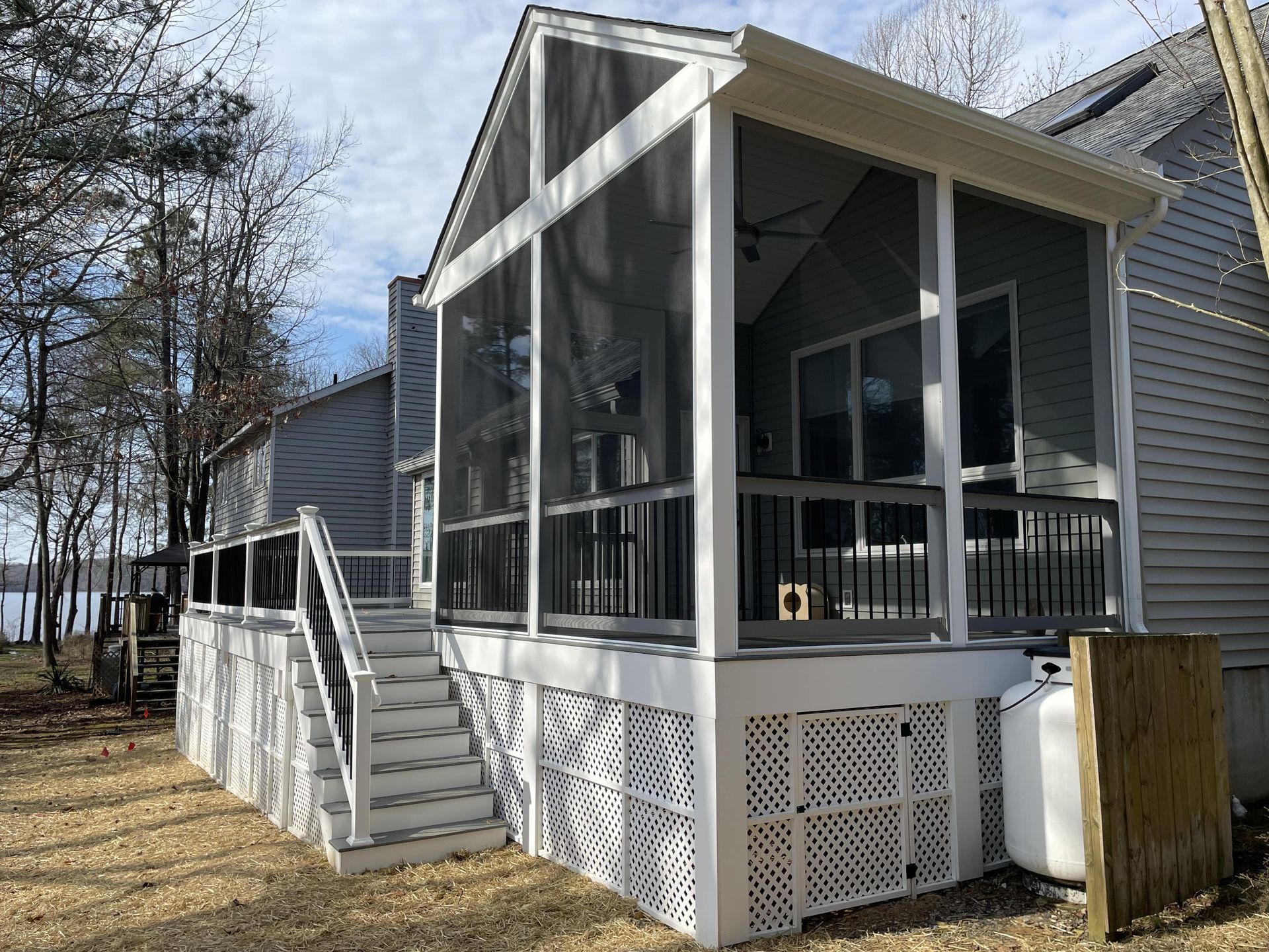 A residential deck builder sanding a wooden deck. Covered front porch with white railing and stairs attached to a gray house in a wooded yard