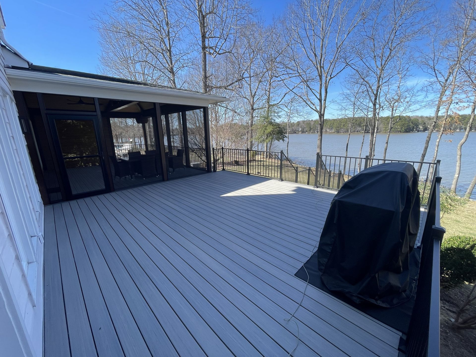 Spacious gray deck beside a lake, with a covered grill and screened porch under a clear blue sky.