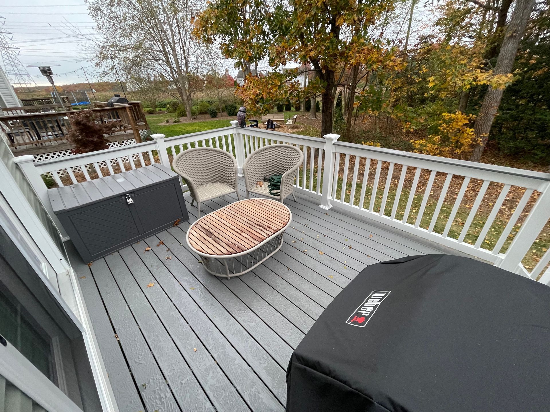 Outdoor deck with gray flooring, white railing, wicker chairs, and a black grill overlooking autumn trees