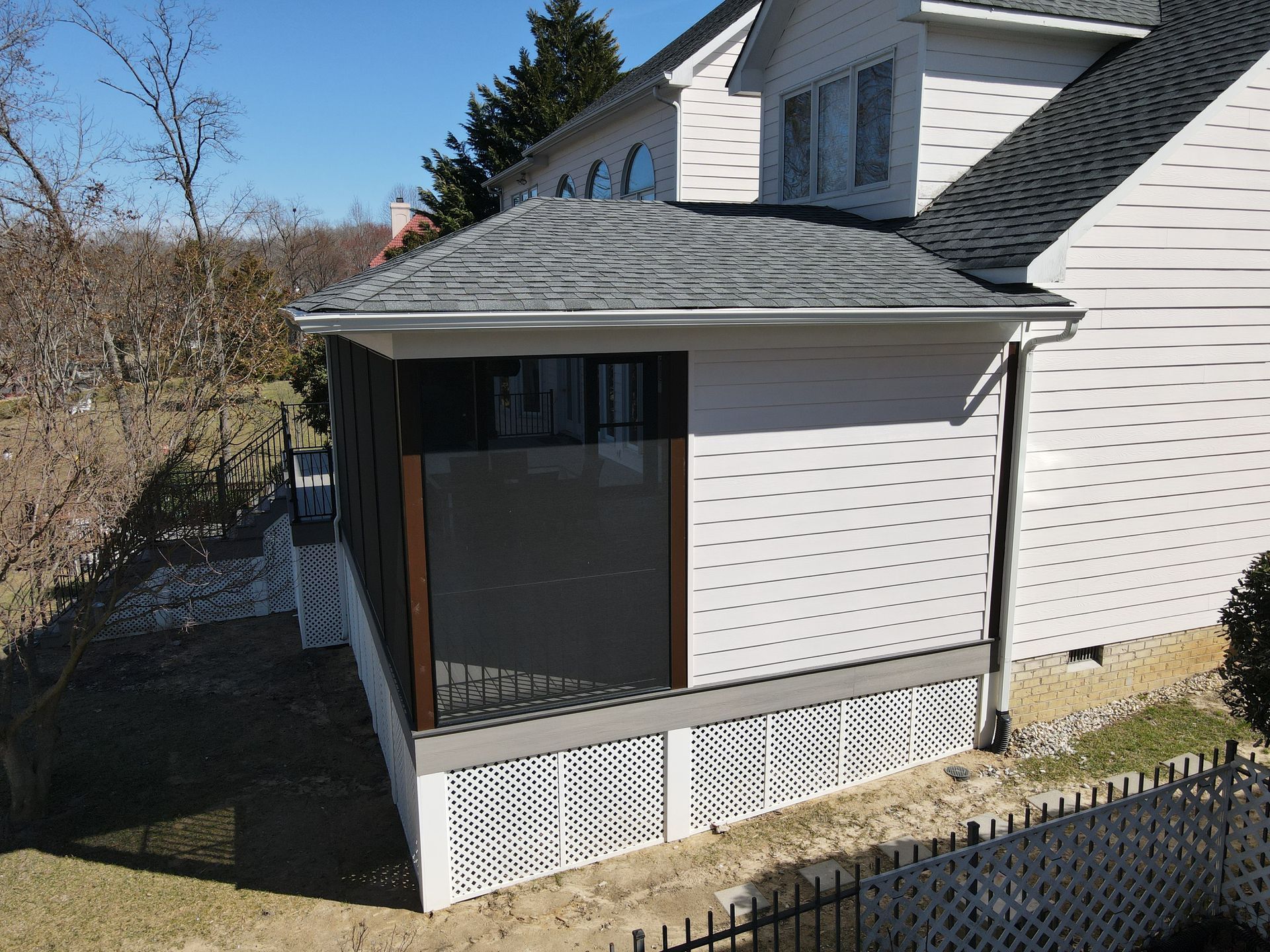 Screened porch on a white house with gray roof and lattice skirting beside a wooded yard.