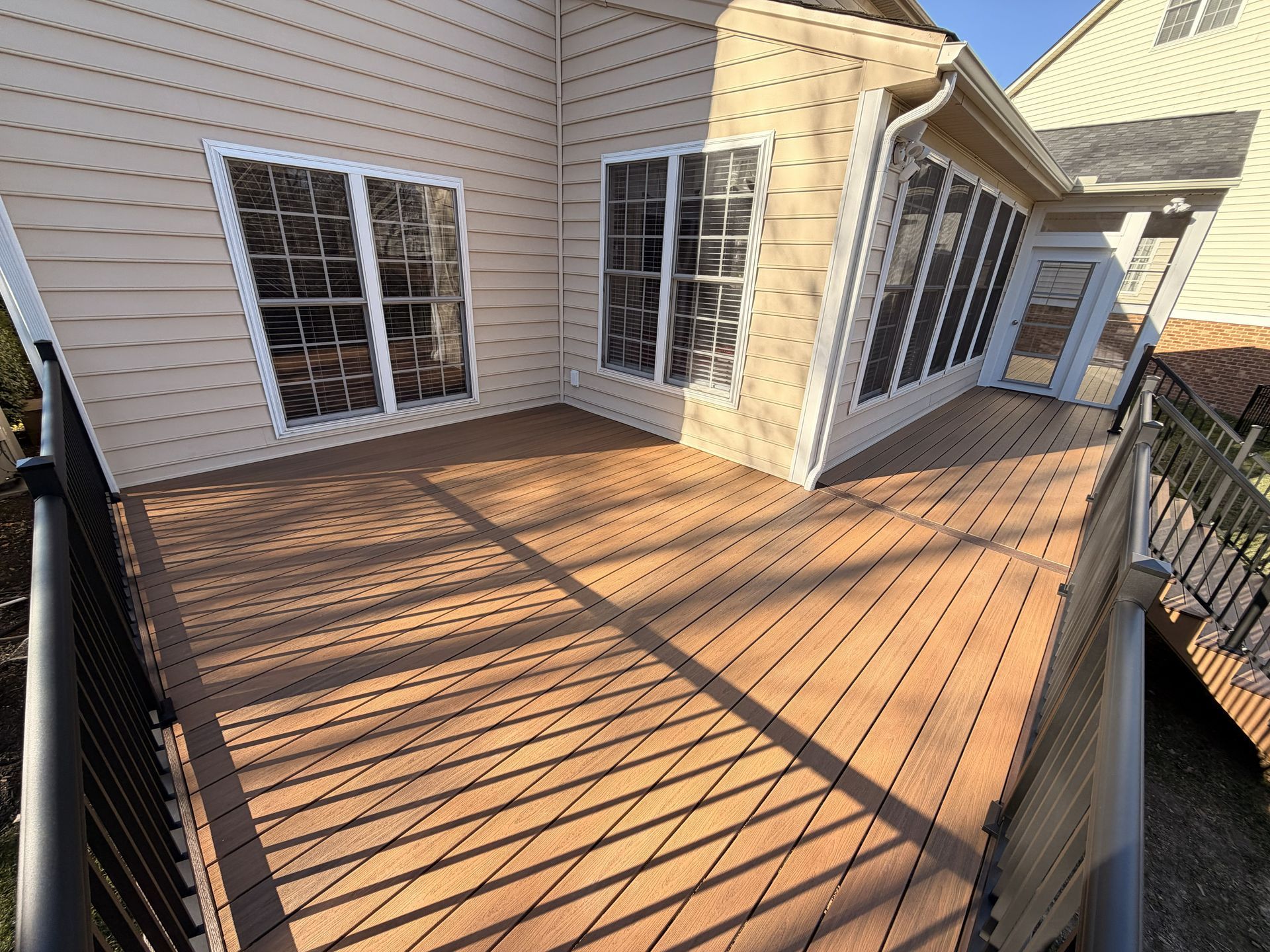 Wooden deck beside beige house with white-trim windows and sloped roof, casting striped shadows