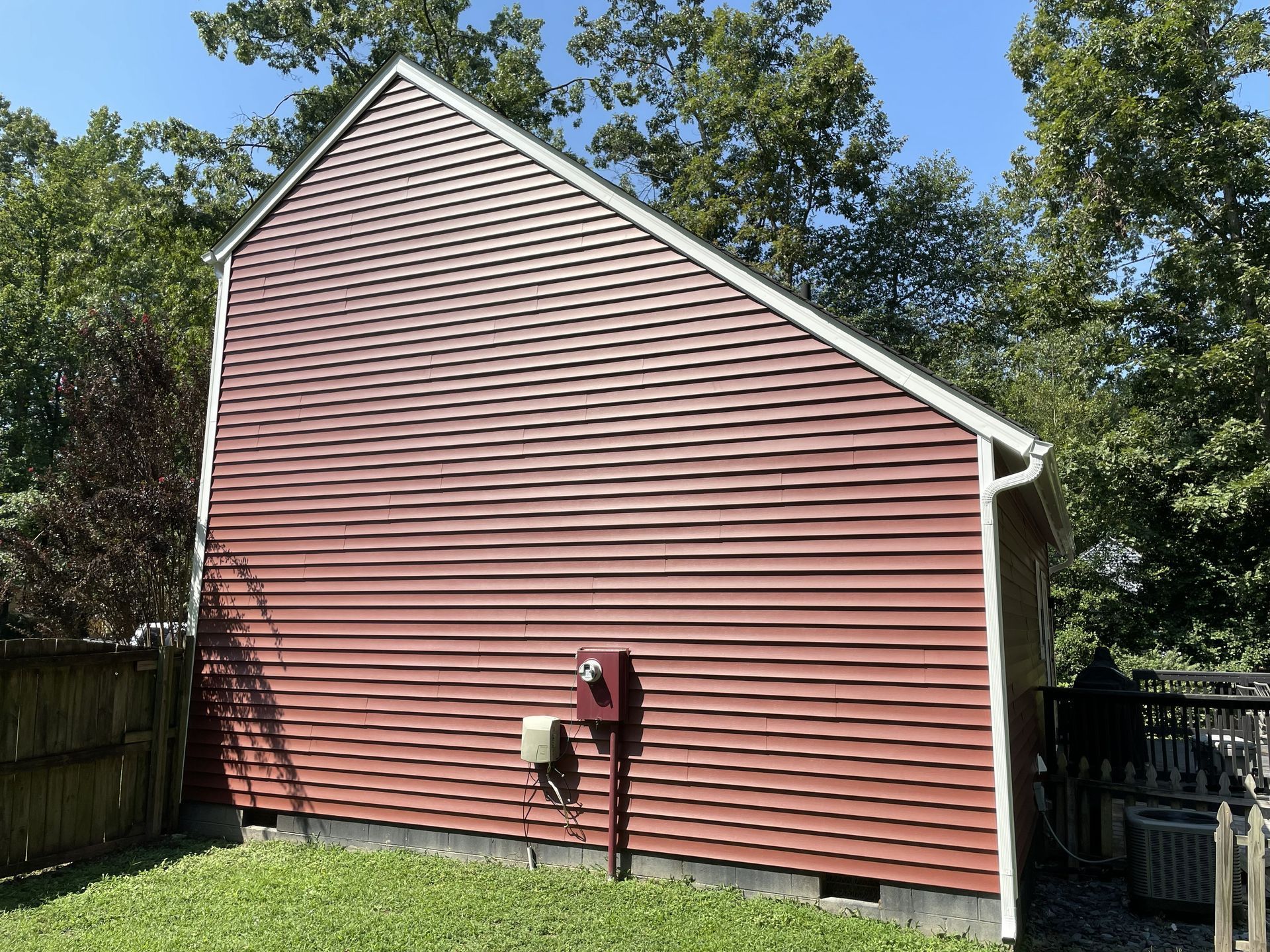 Red-sided house exterior with white trim, utility meter, and fenced yard under trees