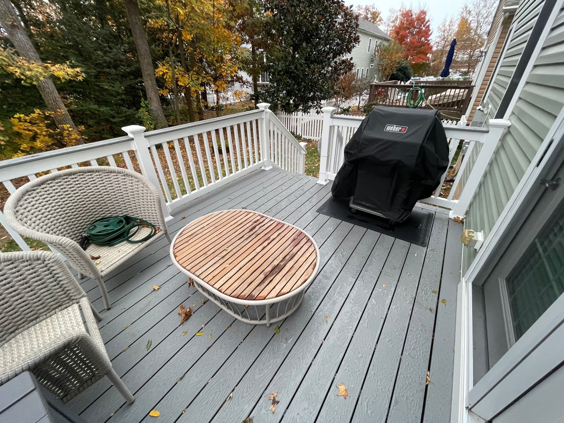 Deck with gray railing, wicker chairs, oval table, and a covered grill overlooking autumn trees.