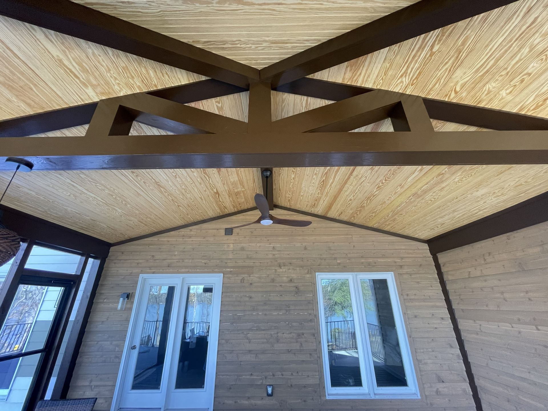 Covered patio ceiling with exposed beams, fan, and windows/door on a brick wall.