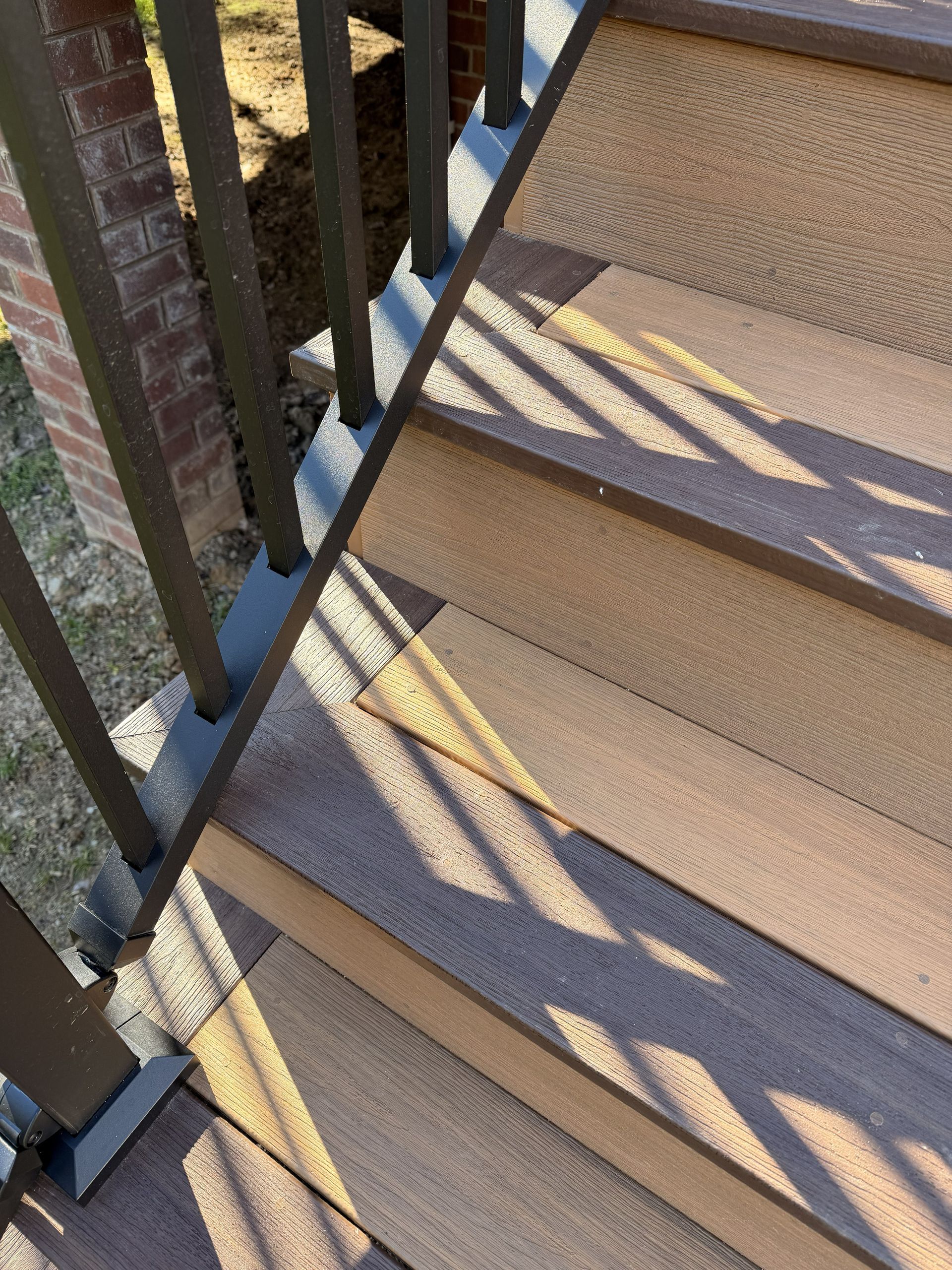 Shadowed outdoor stairs with tan treads and a black metal railing along a brick wall