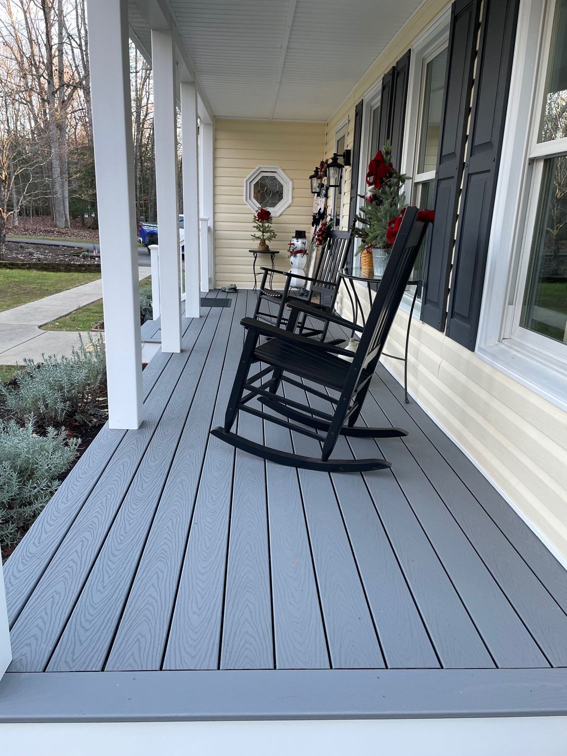 Front porch with gray rocking chair, white columns, and potted flowers along a cream-sided house.