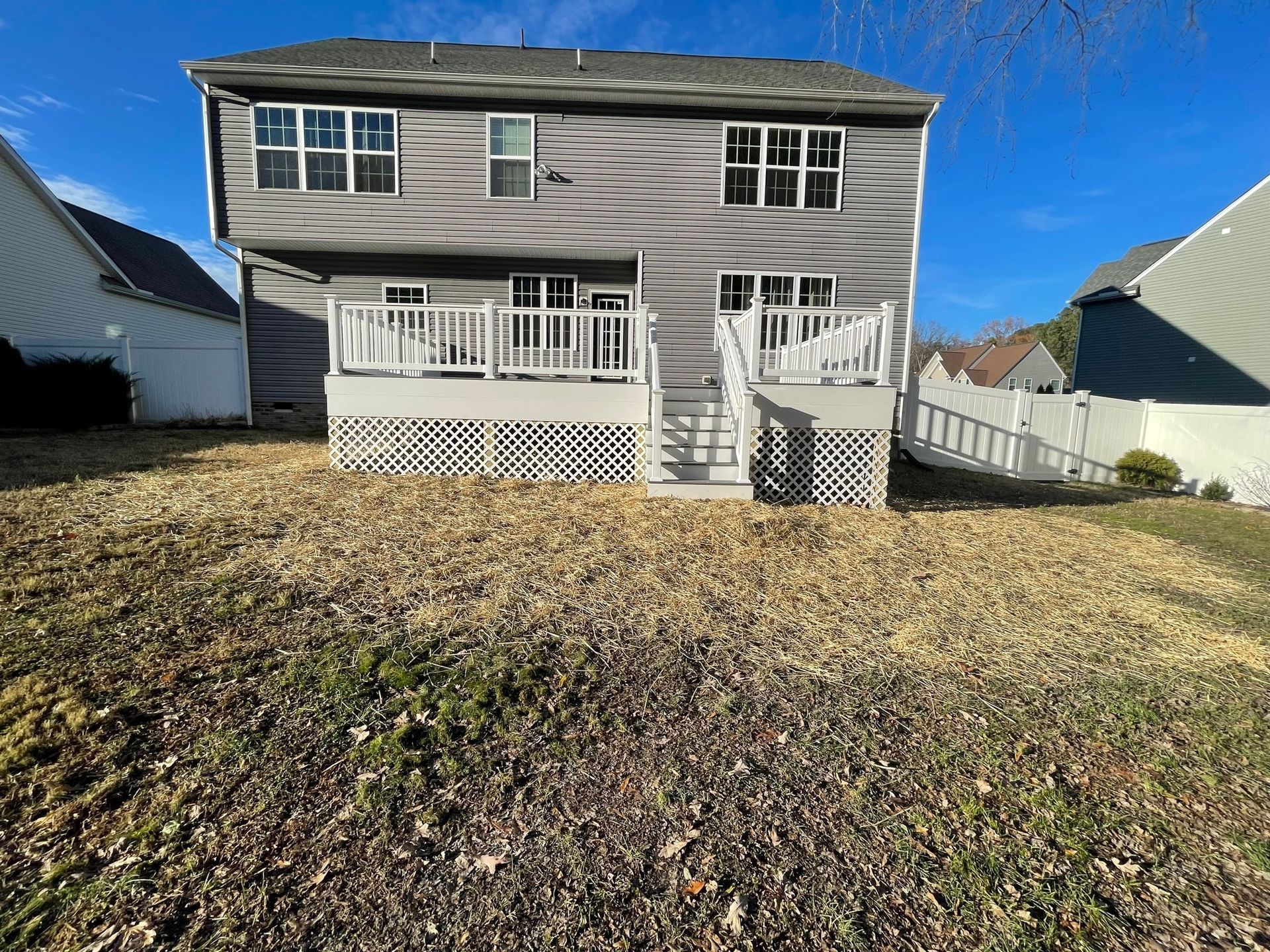 Back of a two-story gray house with a small porch, white trim, and leaf-covered yard in sunlight