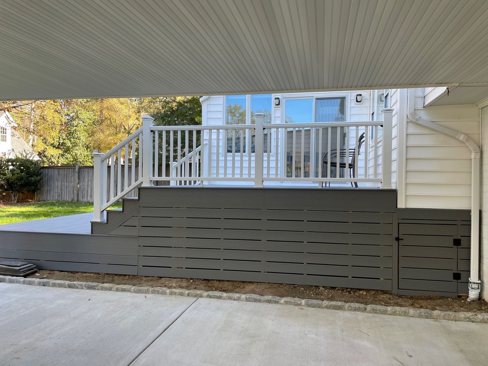 Front porch with white railing, gray skirting, and concrete driveway under a covered overhang.