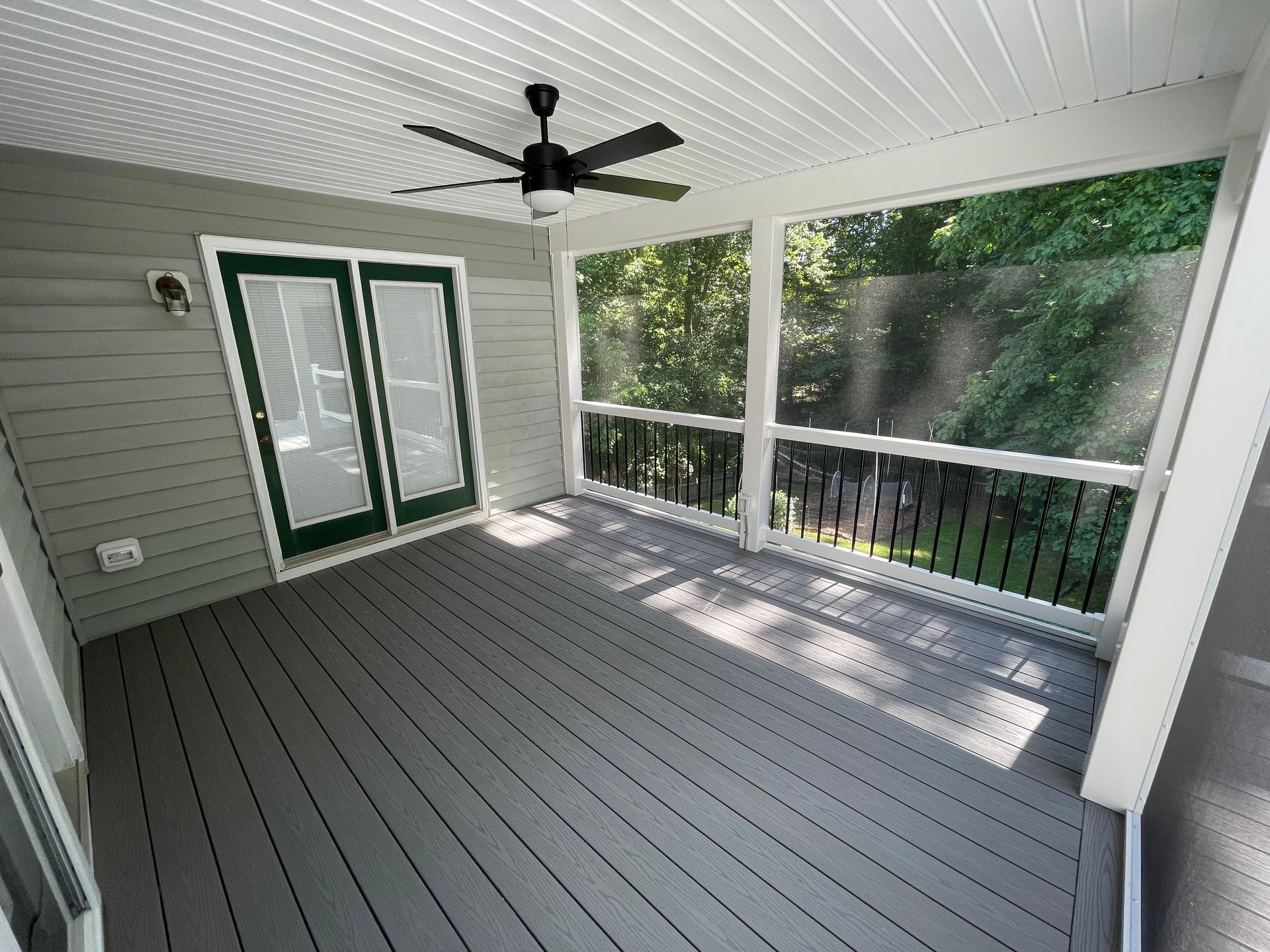 Covered porch with gray flooring, white siding, ceiling fan, and green-trimmed double doors overlooking trees