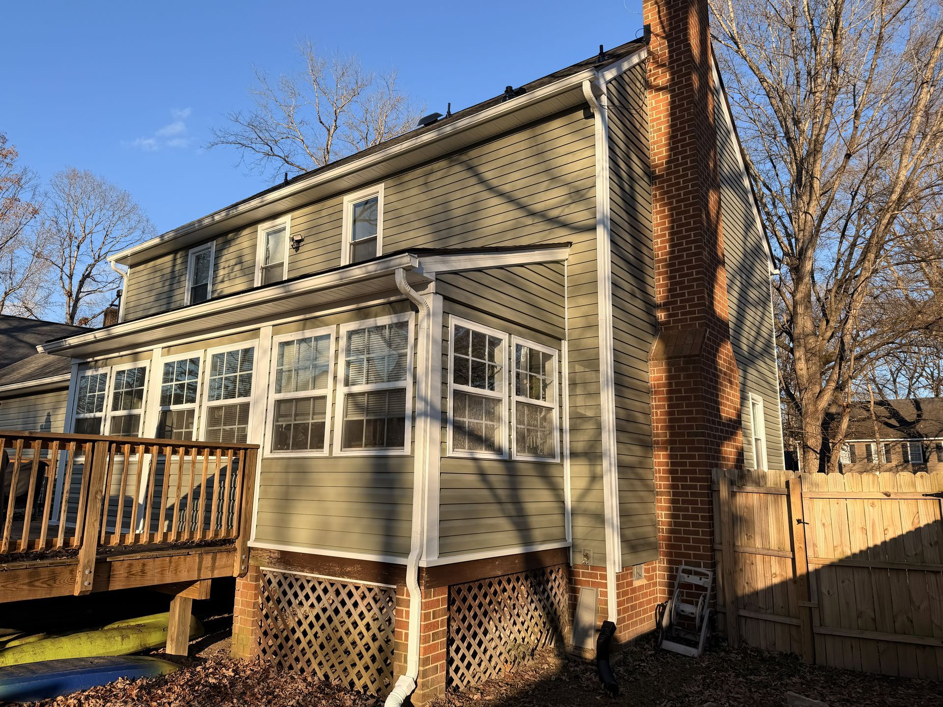 Back of a beige house with a screened porch, wooden deck, brick chimney, and fenced yard at sunset