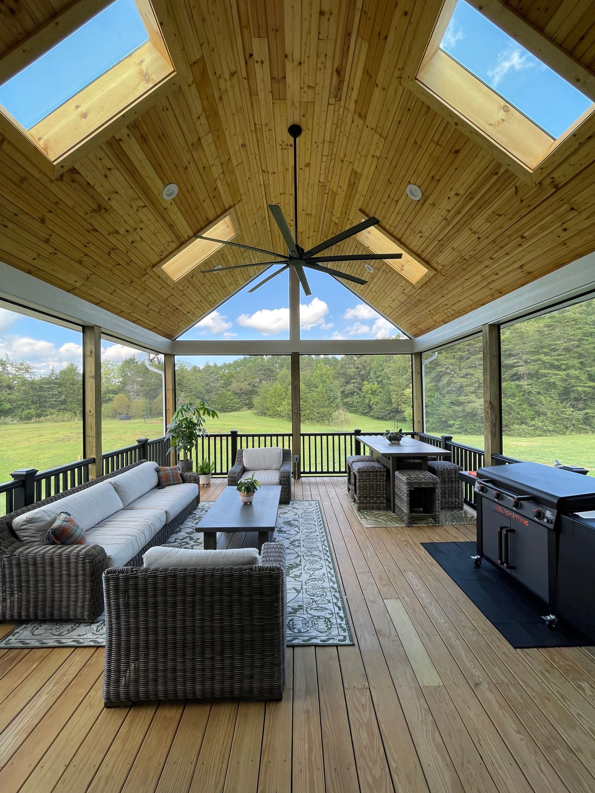 Open-air screened porch with wicker seating, wood ceiling, skylights, and a view of green fields