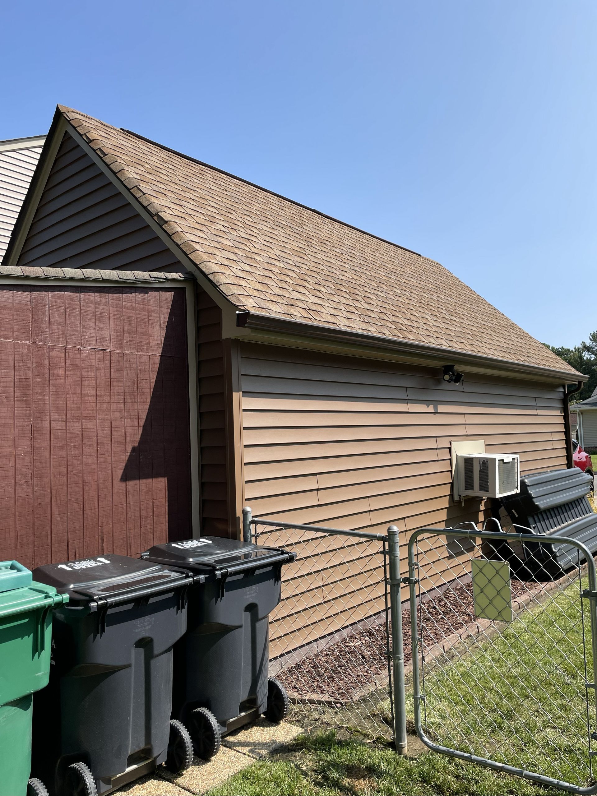 Backyard side of a house with trash bins, chain-link fence, and air conditioner units on the wall