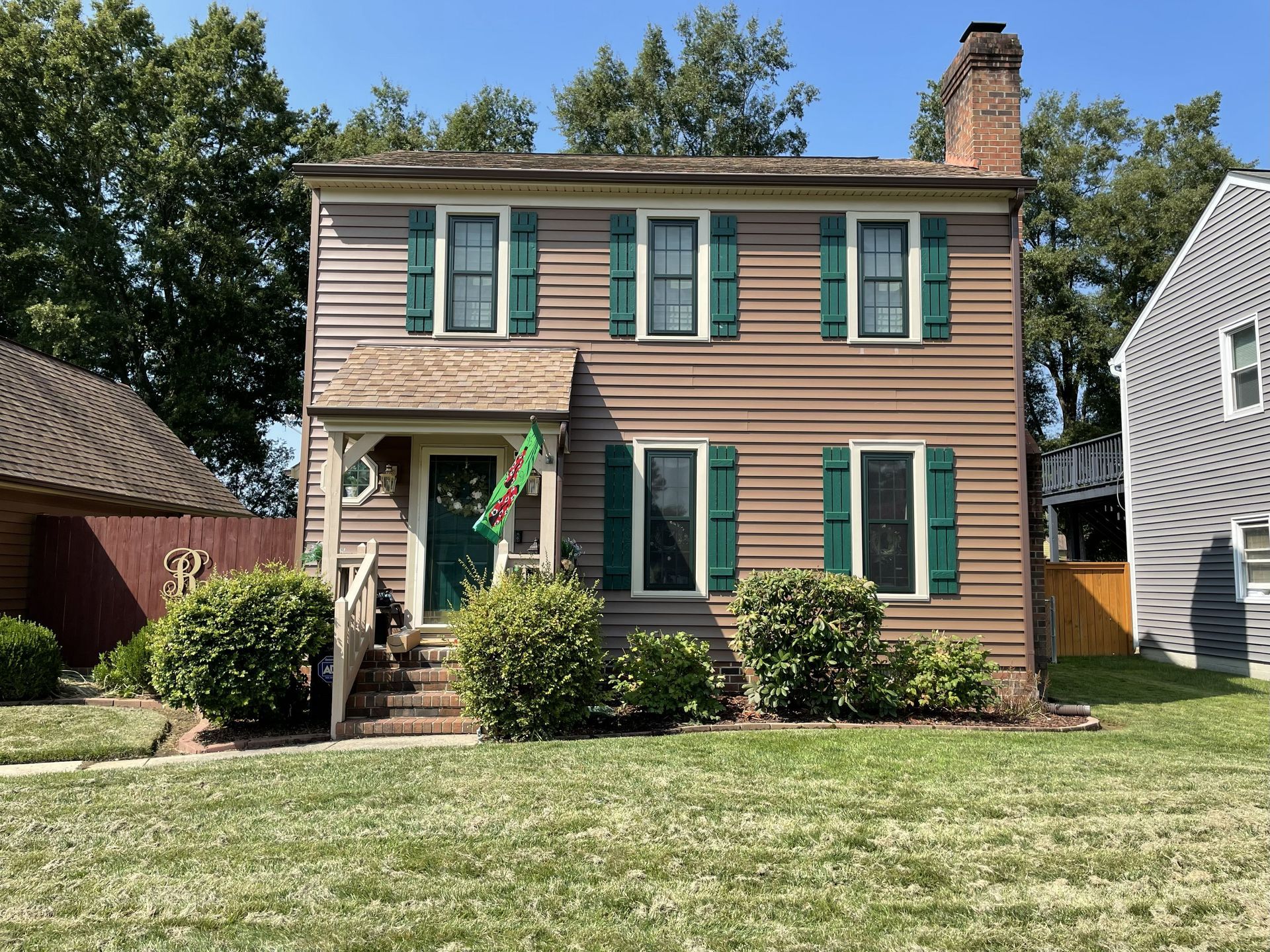 Two-story tan house with green shutters and a front porch, set on a grassy lawn under a blue sky