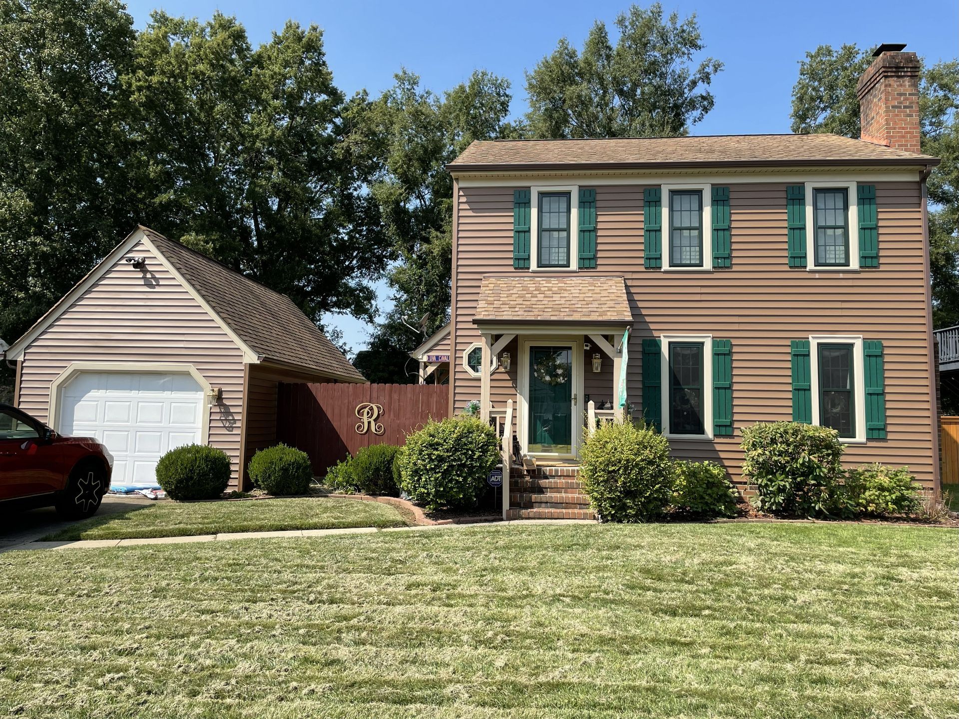 Suburban house with tan siding, green shutters, a detached garage, and a front lawn under a blue sky