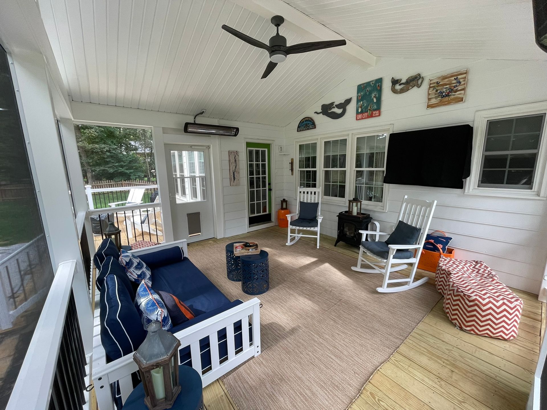 Covered porch with navy sofa, rocking chairs, hanging ceiling fan, and TV on white walls.